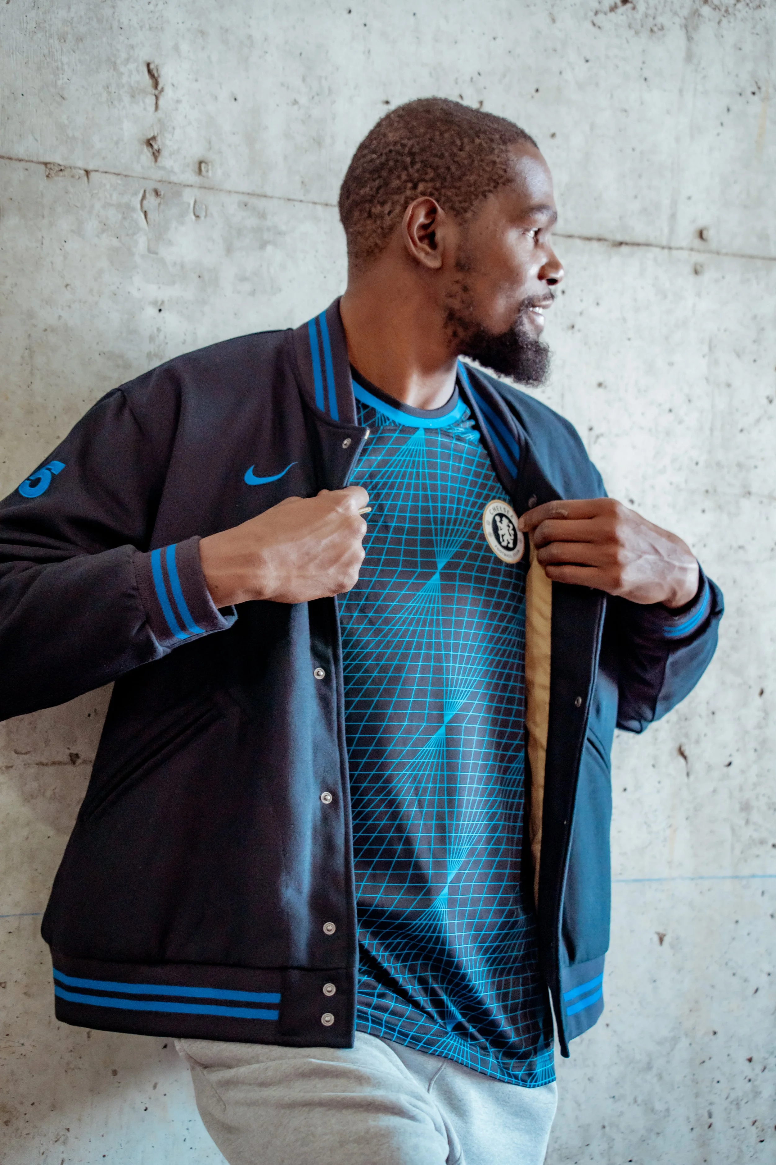 Man in a black and blue Chelsea football jacket opening his jacket to reveal a blue Chelsea jersey featuring a geometric pattern, standing against a concrete wall.