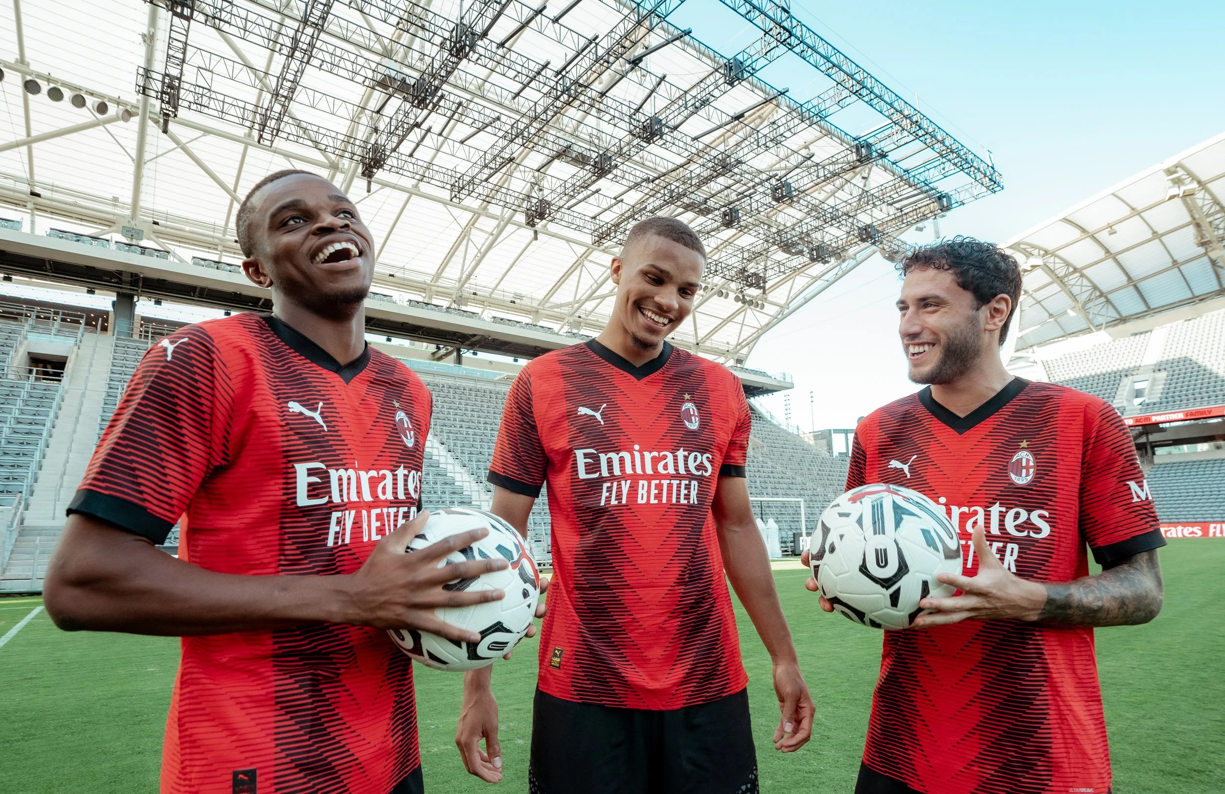 Three male soccer players in red and black uniforms on a soccer field, holding soccer balls, smiling, with a stadium in the background.