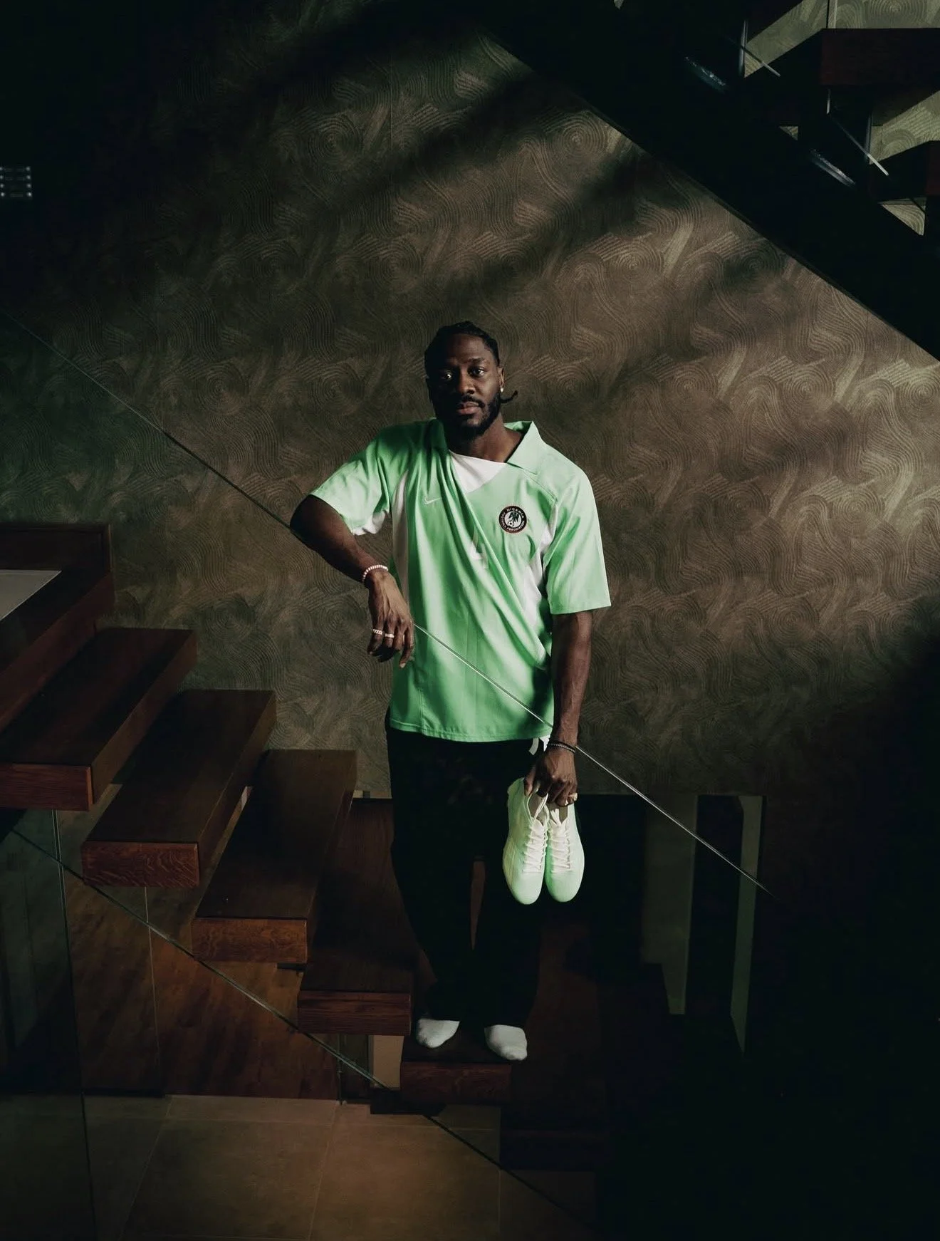 A man standing on a wooden staircase holding a pair of white sneakers, wearing a light green Nike sports jersey with a logo, black pants, and white shoes, against a textured brown wall.