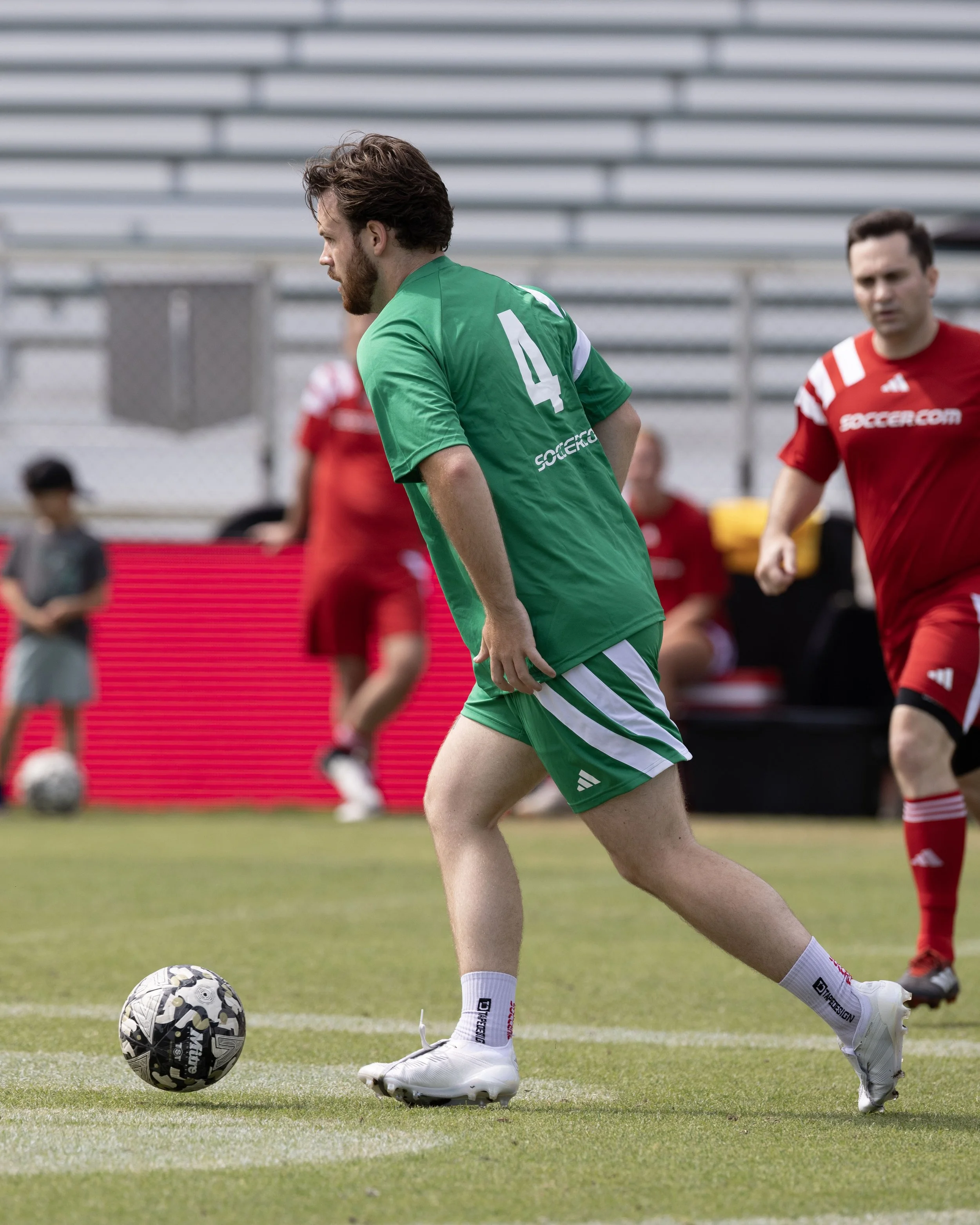 A man wearing a green soccer uniform is kicking a soccer ball on a field during a game. He is surrounded by other players in red uniforms and spectators in the background.