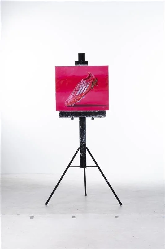 An easel with a small painting of a racing shoe, set against a plain white background.