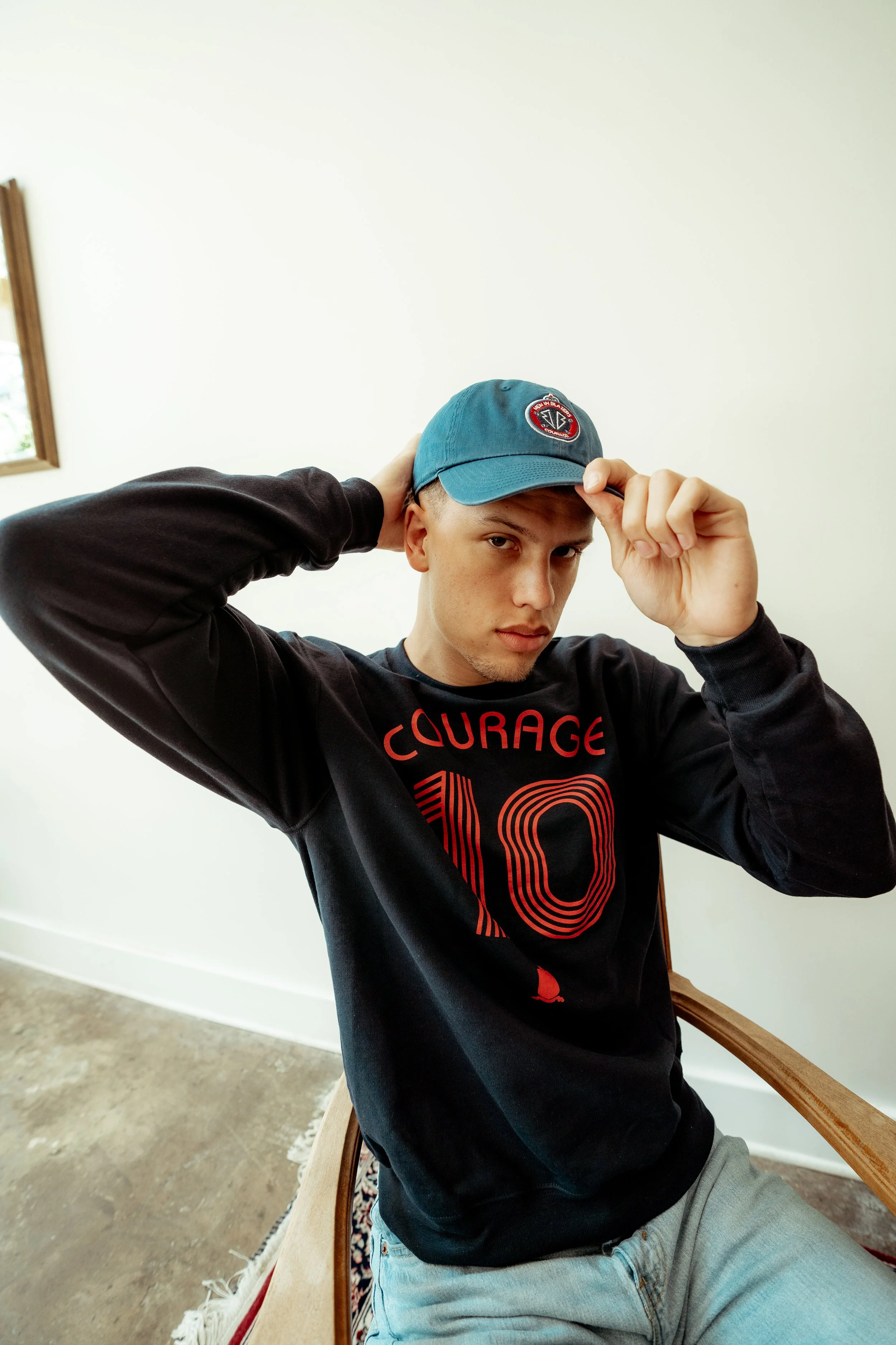 A young man in a black sweatshirt with red writing and a large number 10, wearing a blue cap, sitting on a wooden chair with a white wall in the background.