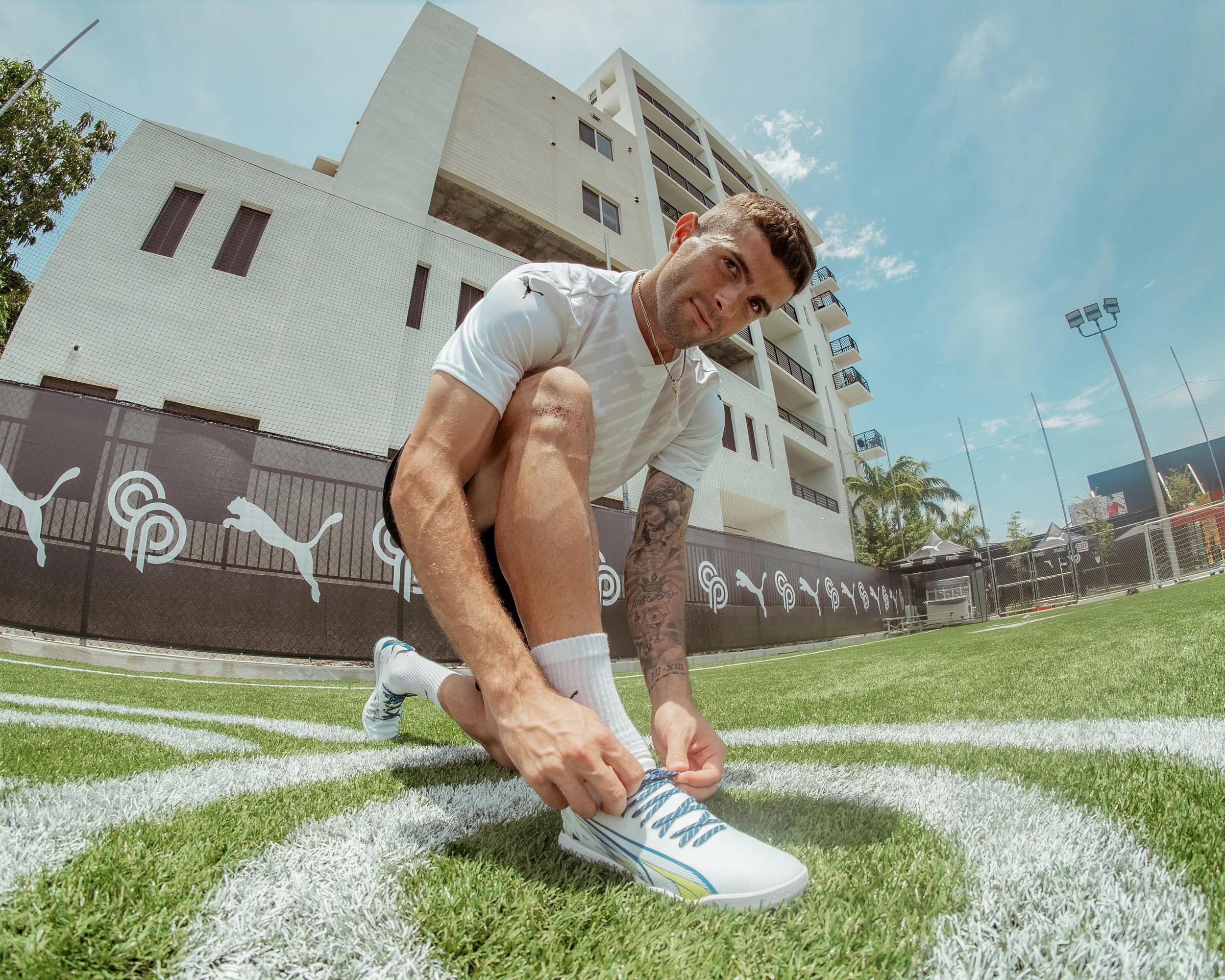 A man in sportswear tying the laces of his soccer cleats on a soccer field, with apartment buildings and a fence in the background.