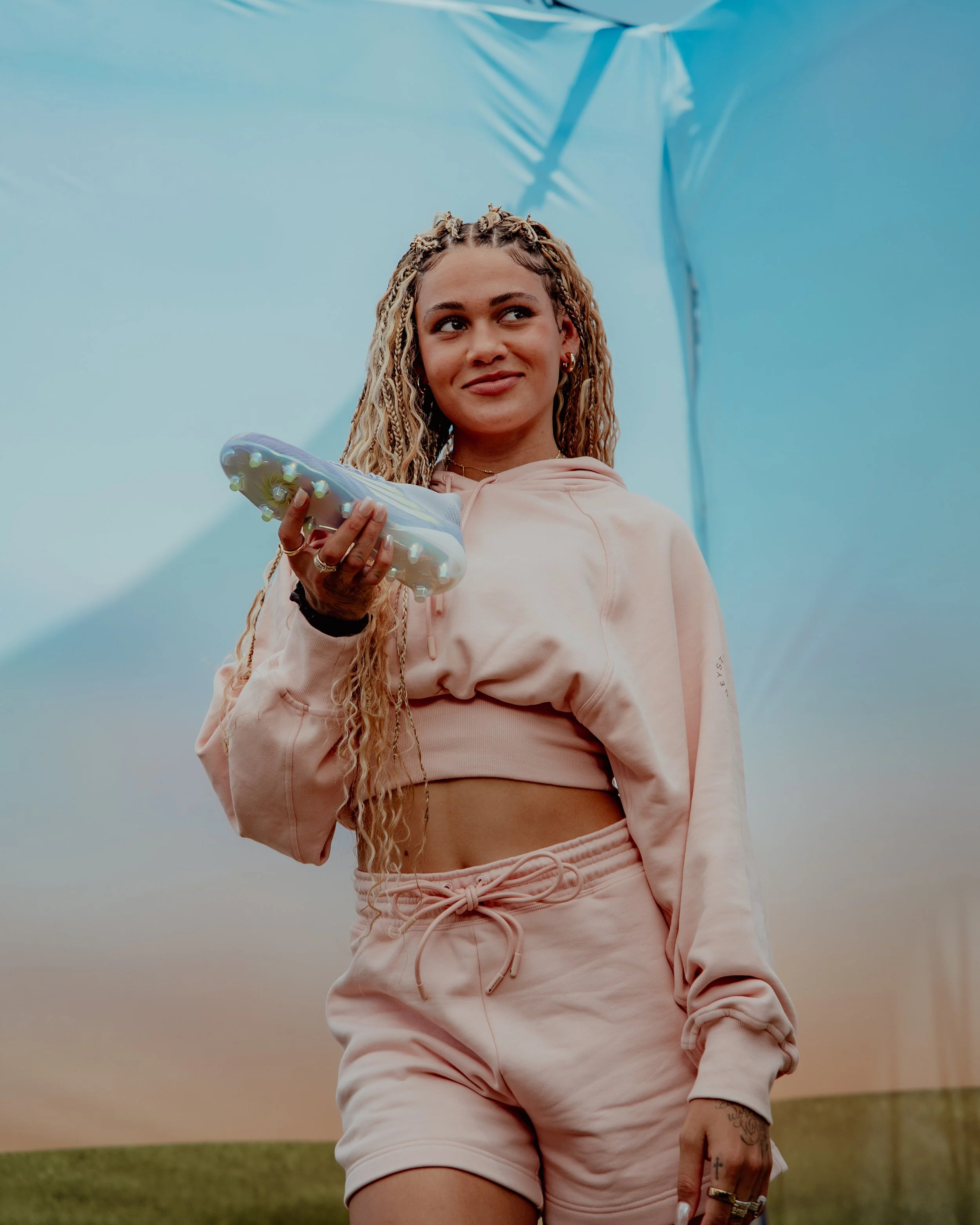 A young woman with braided blonde hair holding a light-colored athletic shoe, wearing a beige cropped hoodie and matching shorts, standing outdoors against a blue tent background.