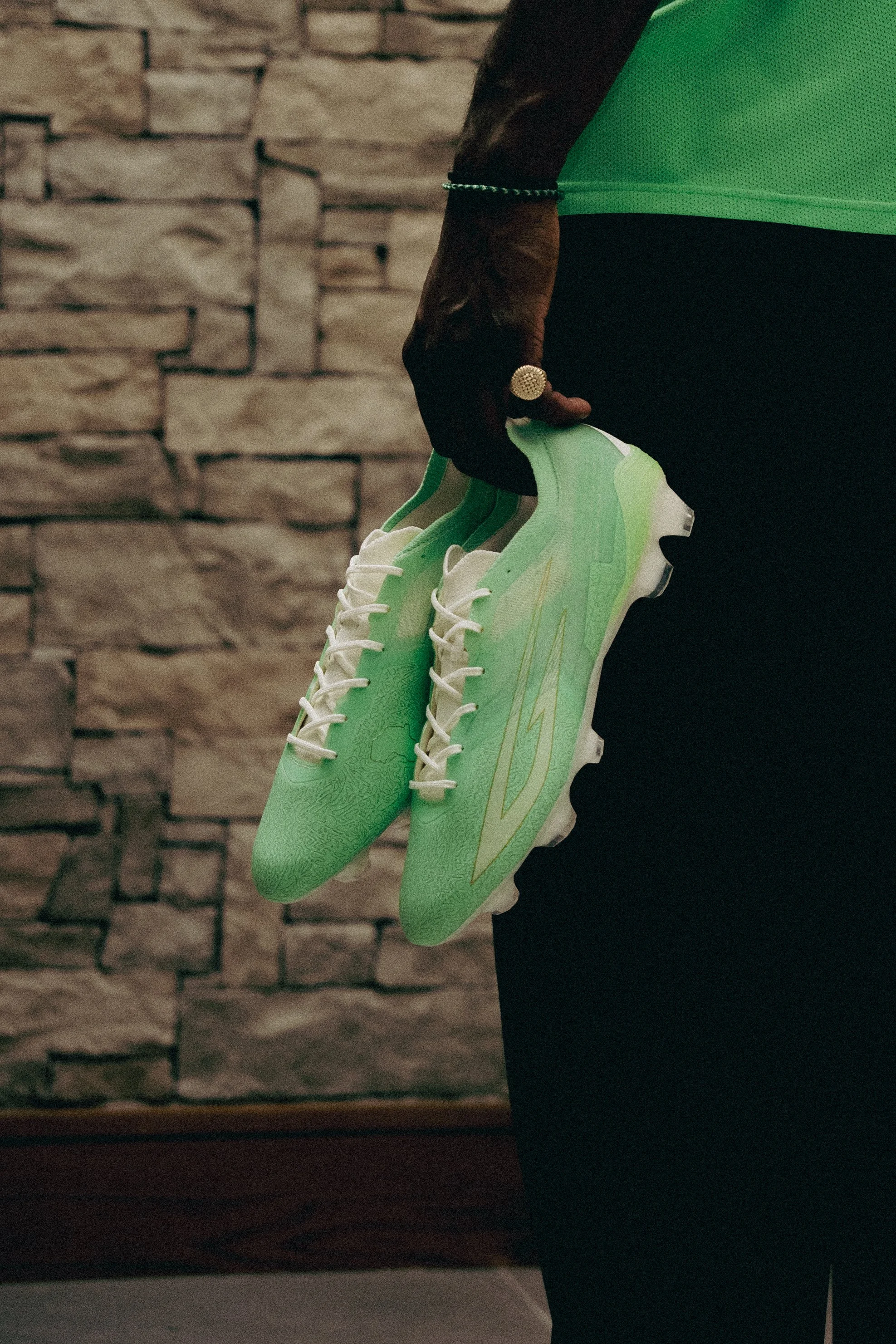 A person holding a pair of light green athletic shoes with white soles and laces, against a background of a brick wall.