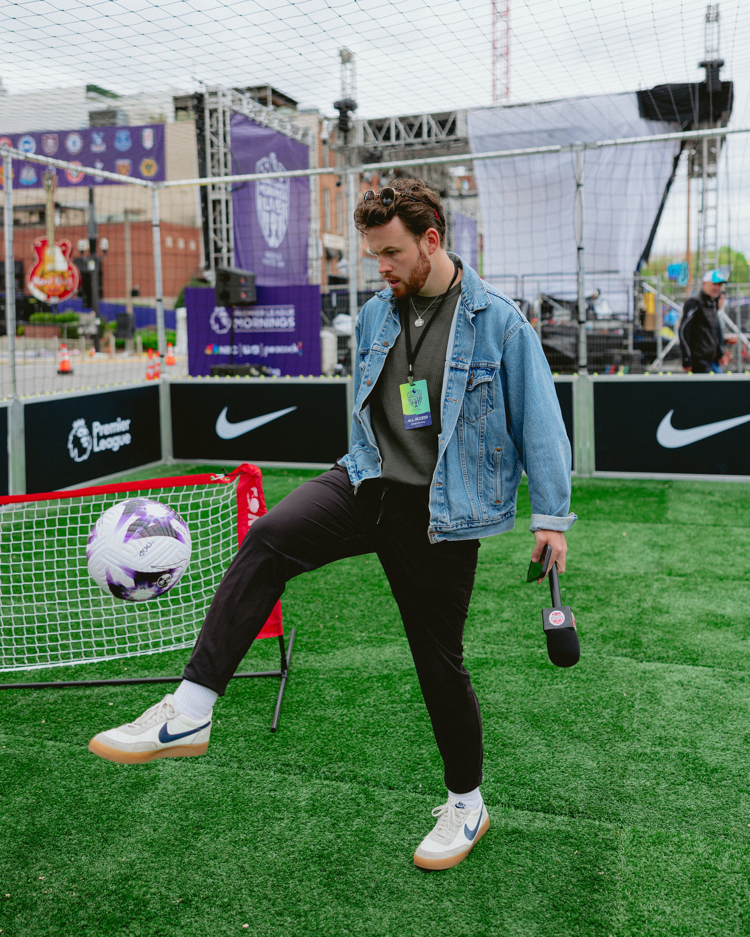 Young man in a denim jacket kicking a soccer ball during a sports event, holding a microphone, on a green field with banners and stage in the background.