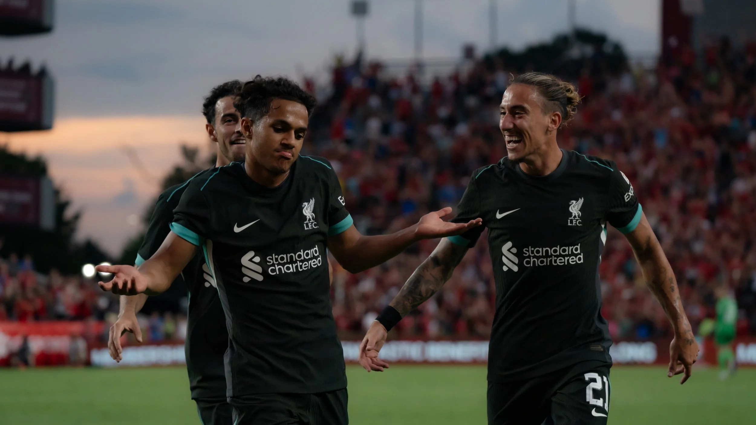 Soccer players from Liverpool Football Club celebrating on the field during a match, wearing black jerseys with the nike logo and standard chartered sponsorship, against a backdrop of a cheering crowd.