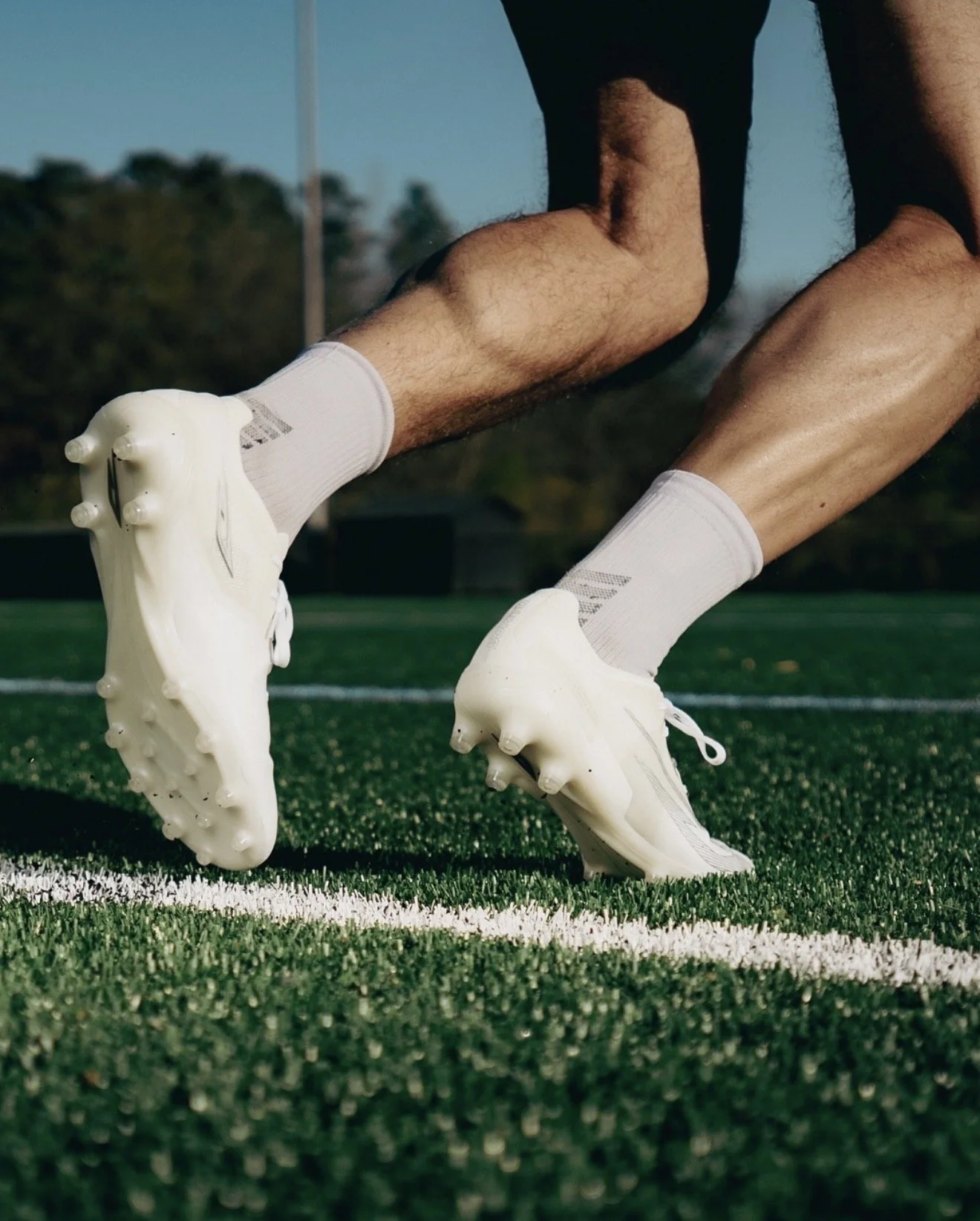 Close-up of a person wearing white sports shoes and white socks, crouched on an outdoor football field with green artificial turf, preparing to run or jump.