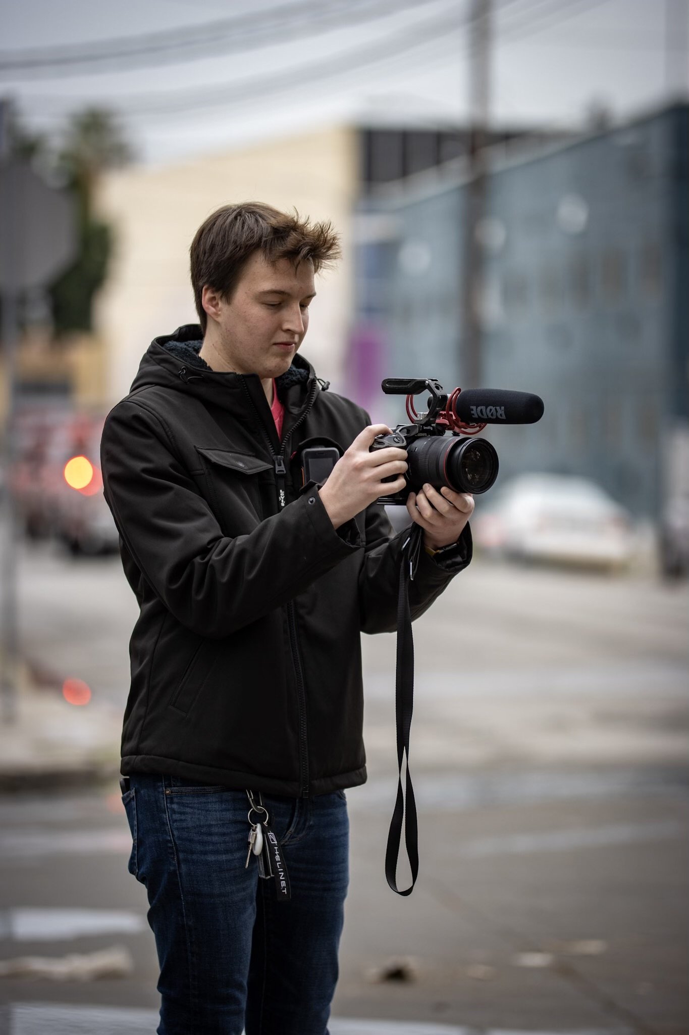 Person holding a camera with a microphone attached, standing outdoors in a casual jacket and jeans.