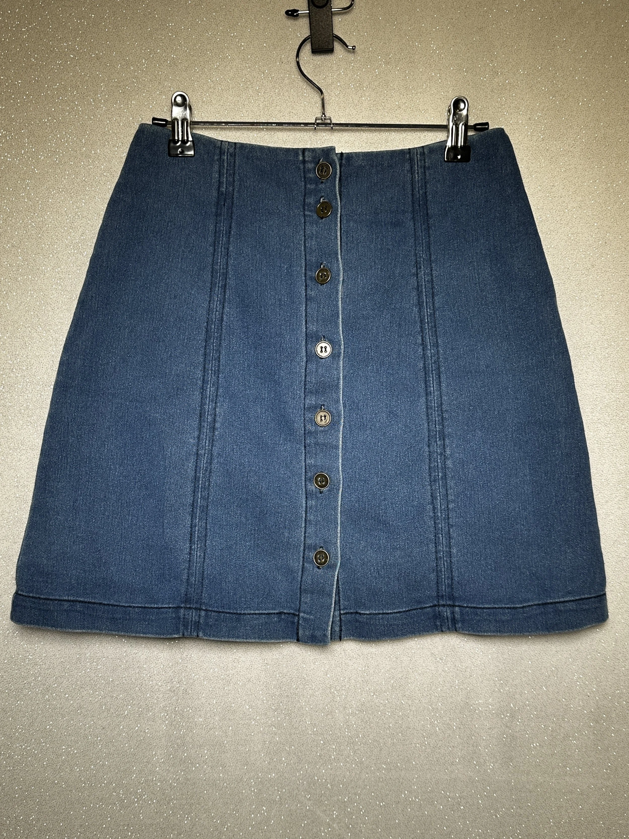 Blue denim skirt with front buttons hanging on a hanger against a glittery wall.