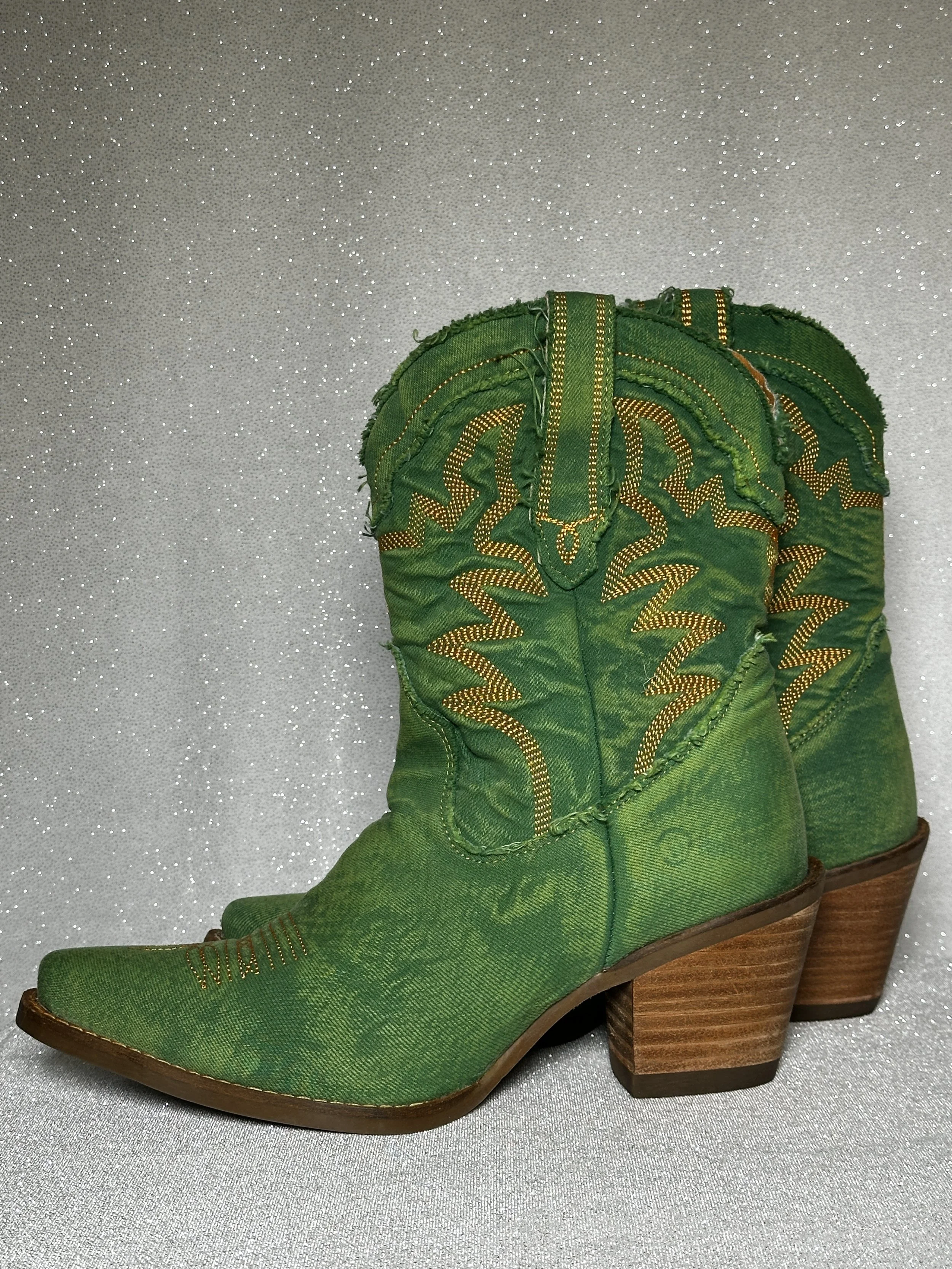 A pair of green cowboy boots with decorative yellow stitching, pointed toes, and stacked wooden heels, displayed against a glittery silver background.