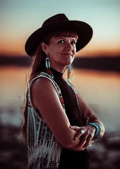 A woman with long hair, wearing a black hat, turquoise earrings, and a patterned vest, standing outdoors during sunset.