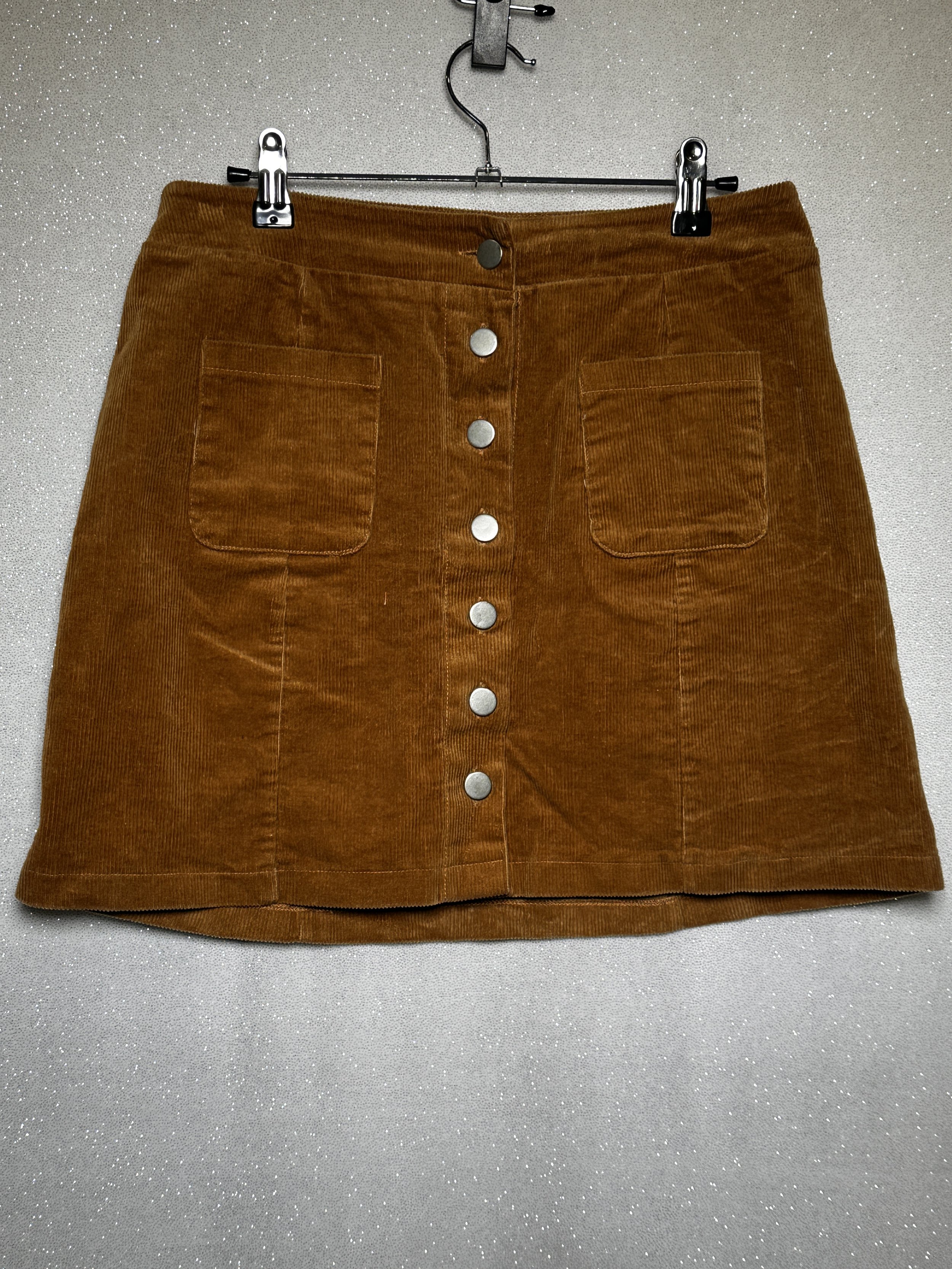 Brown corduroy skirt with front buttons and patch pockets hanging on a clothes hanger against a sparkly background.