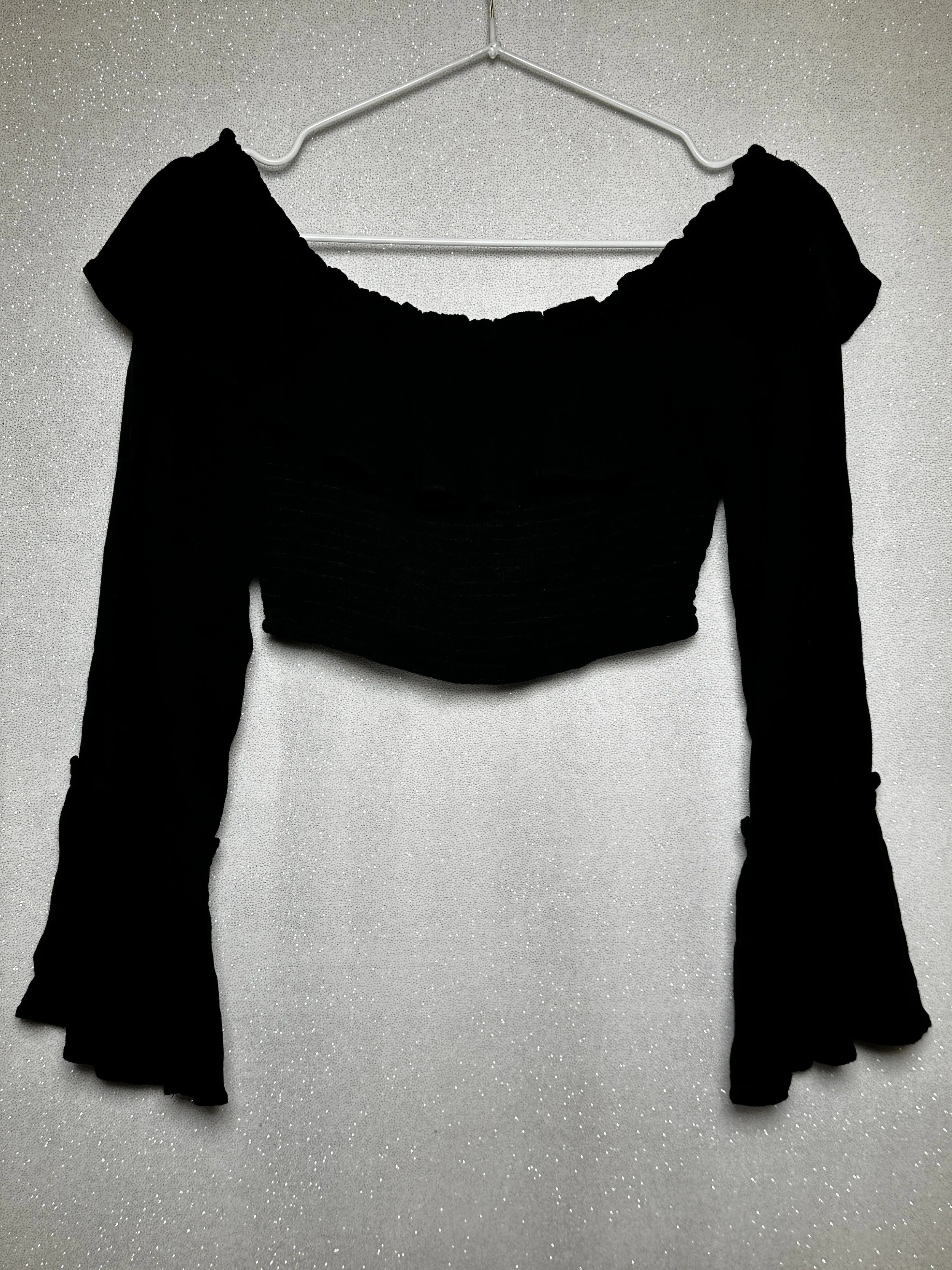 Black off-the-shoulder long sleeve top hanging on a wire hanger against a silver glittery background.