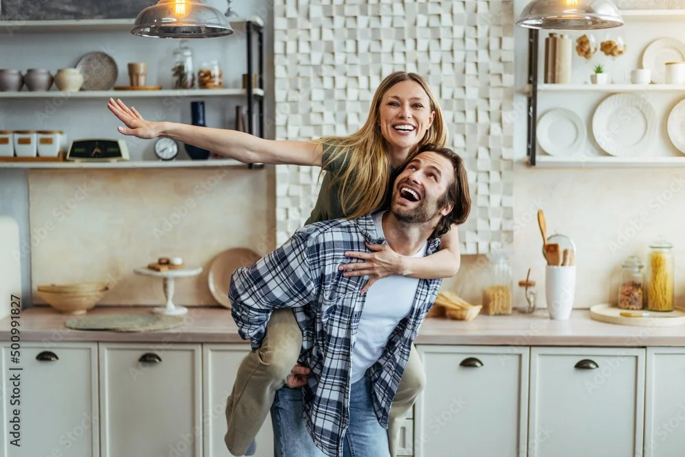 A woman giving a piggyback ride to a man in a kitchen, both smiling and looking happy.