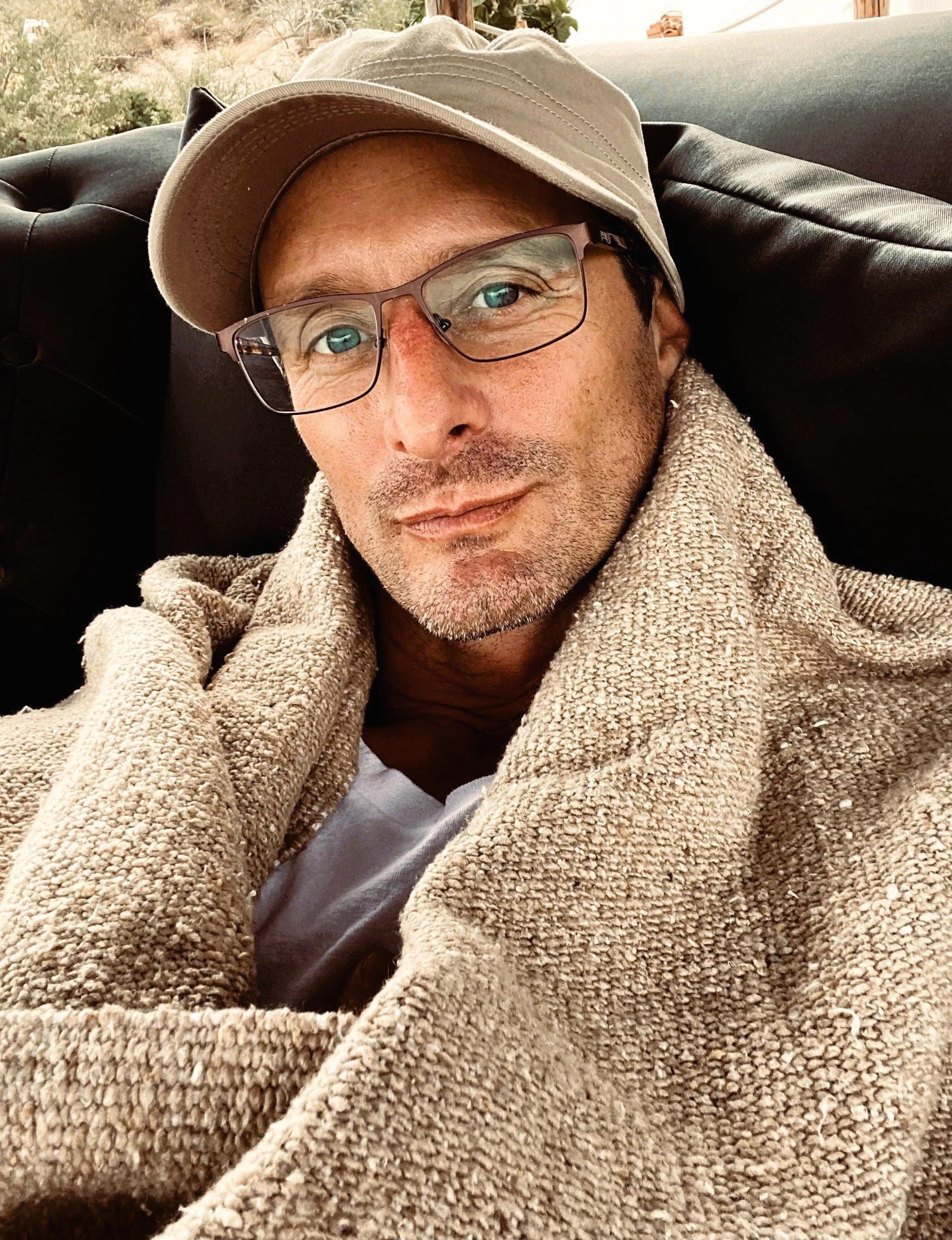 A man with glasses, a beige baseball cap, and a tan textured blanket around his shoulders, resting on a black leather piece of furniture with a desert landscape view in the background.