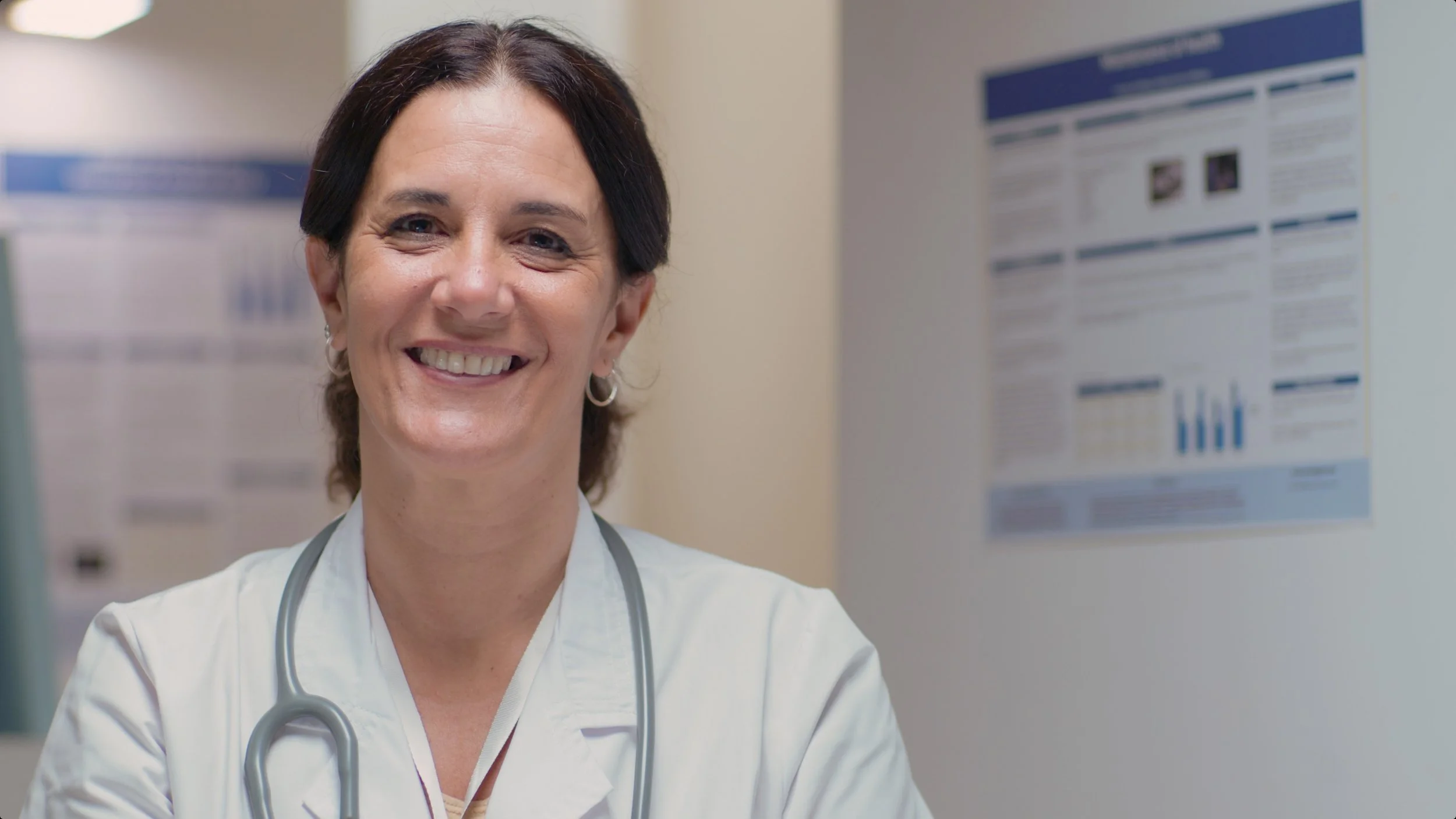 A smiling female healthcare professional wearing a white coat and stethoscope in a clinical setting.