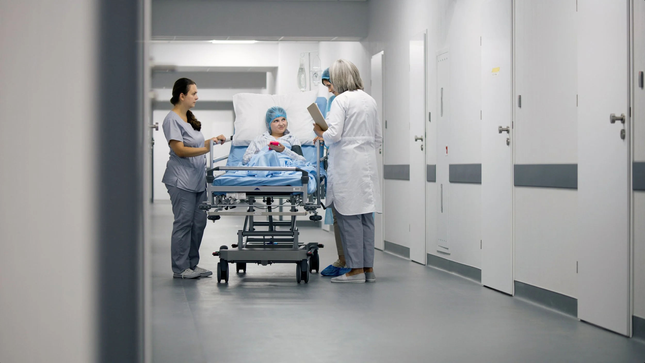 A young girl in a hospital bed, wearing a blue cap, is being visited by a female nurse, a female healthcare worker, and an older female doctor in a hospital corridor.