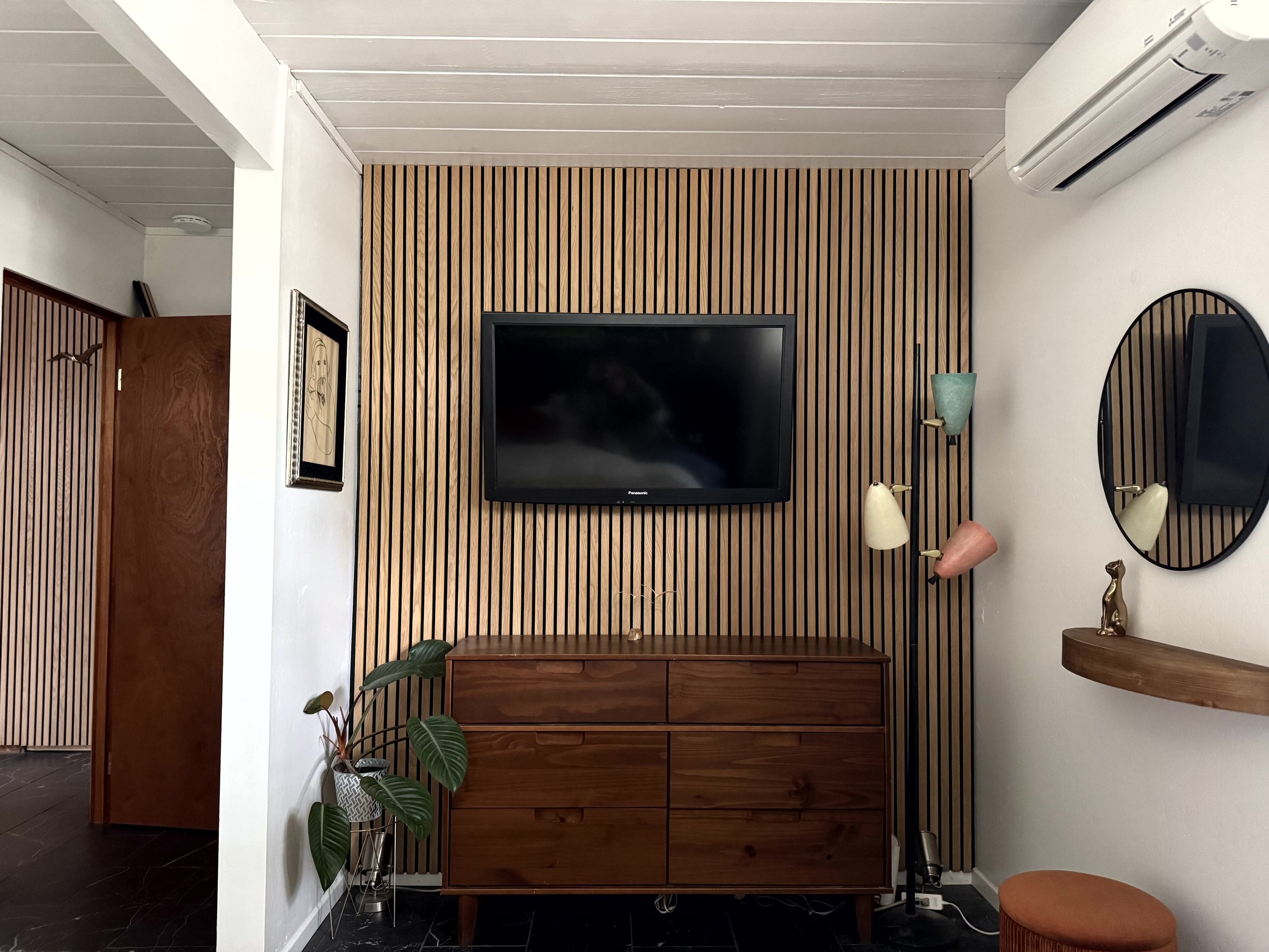 Living room with wooden slat accent wall, wall-mounted flat screen TV, round mirror, floor lamp with four colorful shades, wooden sideboard, potted plant, and an air conditioner.