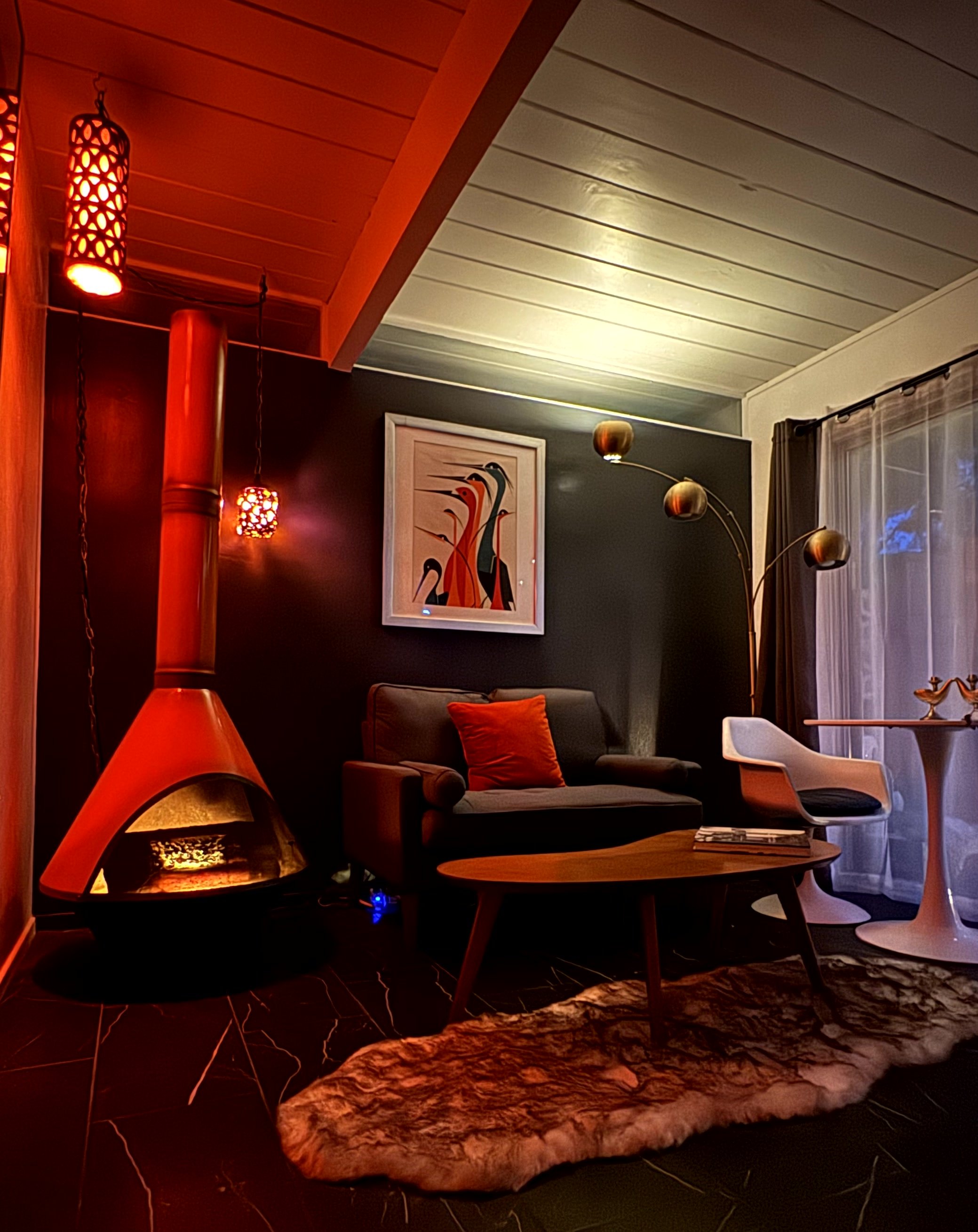 Cozy living room with a black wall, mid-century modern sofa with a red pillow, wooden coffee table, white armchair, unique multi-armed floor lamp, colorful abstract artwork on the wall, red pendant lights, and a Sheepskin rug on a dark tiled floor ne