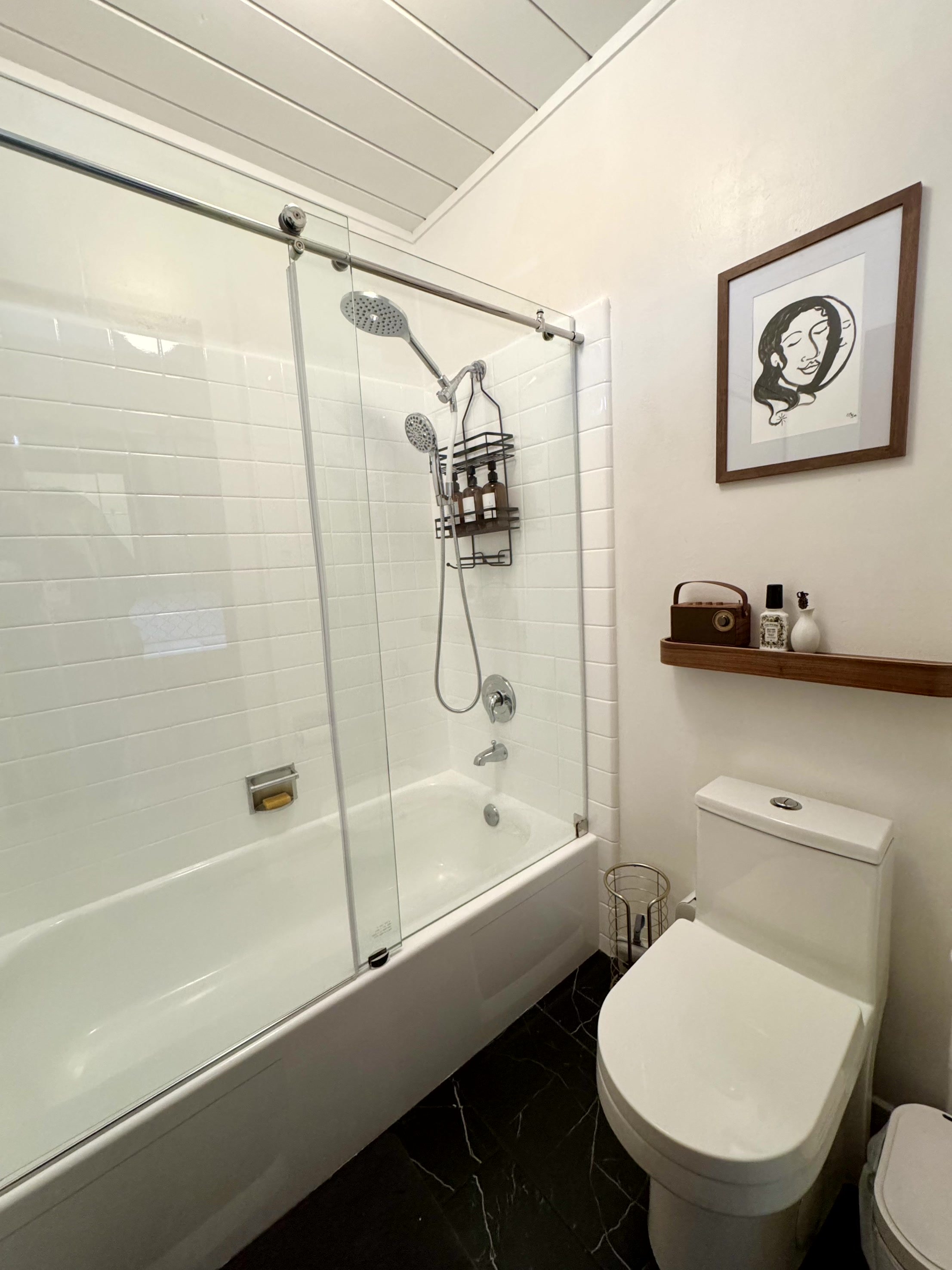 A bathroom with a bathtub, shower area with glass sliding door, wall-mounted shower heads, a wall shelf with toiletries, a framed art piece on the wall, a toilet with a wood shelf above, and black marbled flooring.
