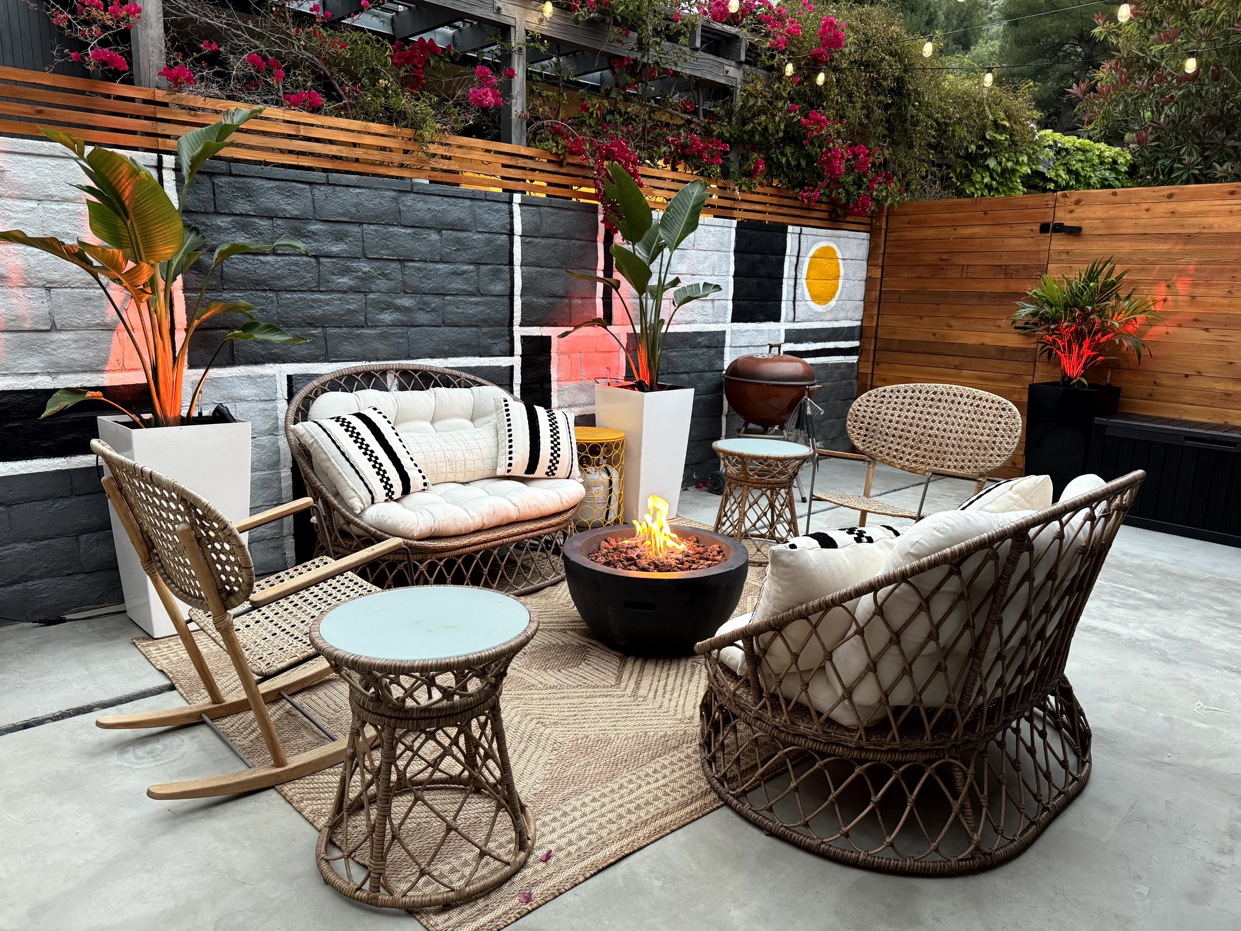 A cozy outdoor patio with wicker furniture, a fire pit, and vibrant plants at dusk, featuring a mural on a black brick wall and string lights overhead.