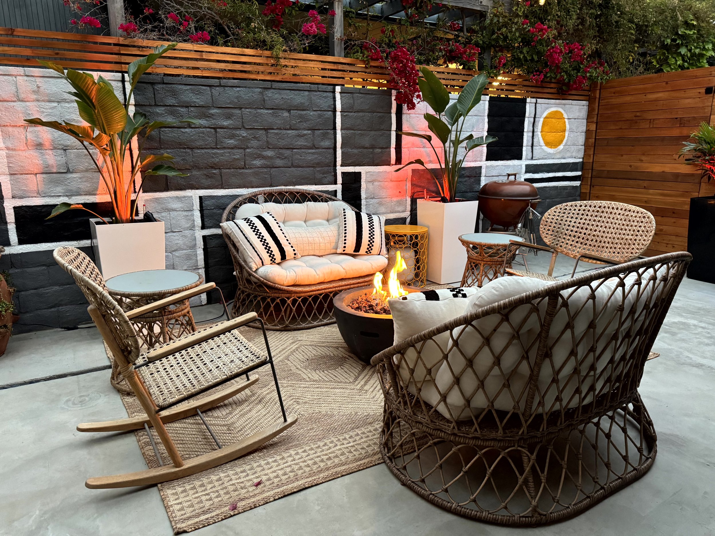 Outdoor patio with wicker and rattan furniture, a small fire pit, potted plants, a decorative wall painting, and a wooden privacy fence.