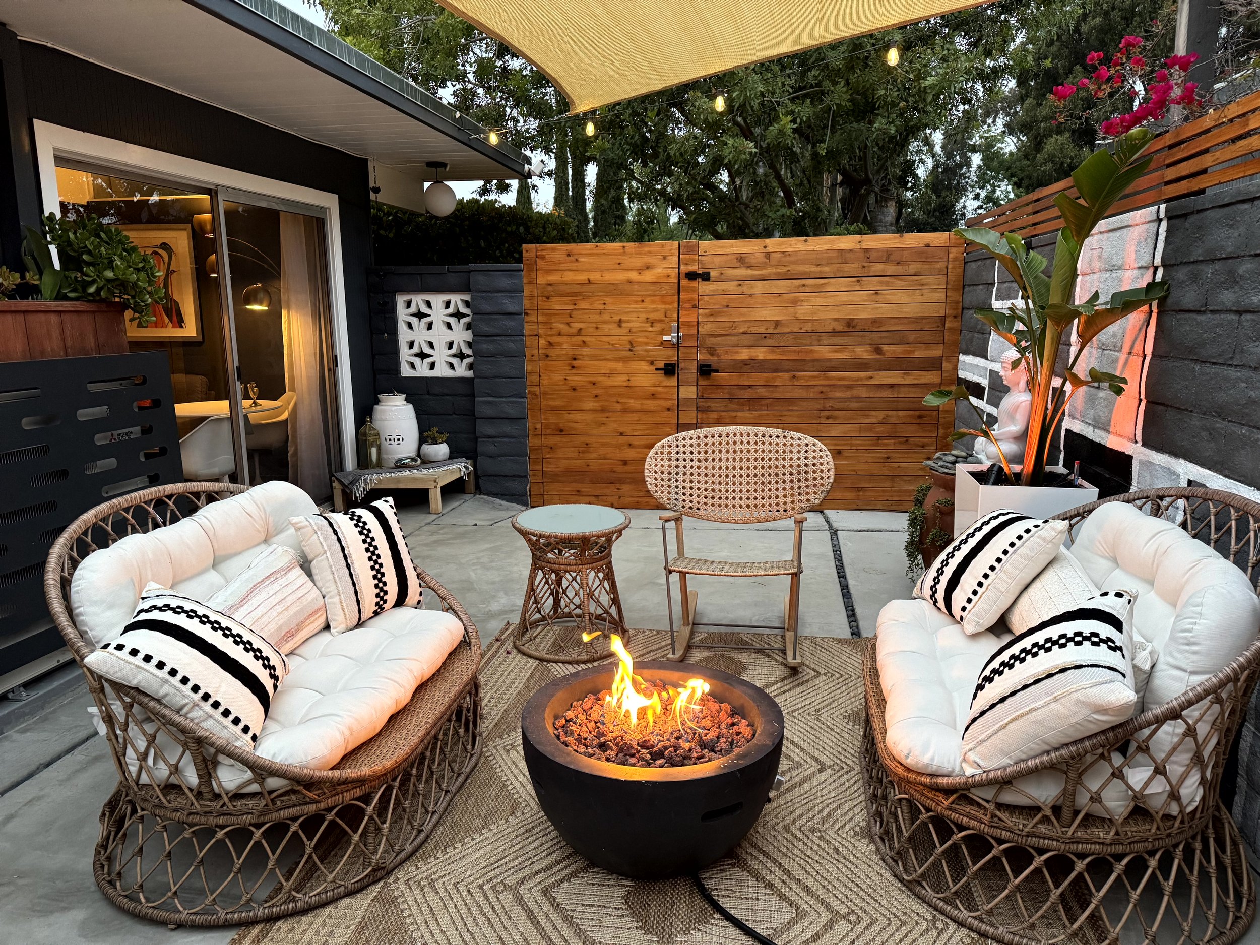 Outdoor patio with two cushioned wicker loveseats, a small round wicker table, and a fire pit in the center, surrounded by decorative pillows. There are potted plants, a wooden privacy fence, and string lights overhead. A doorway leads into an interi