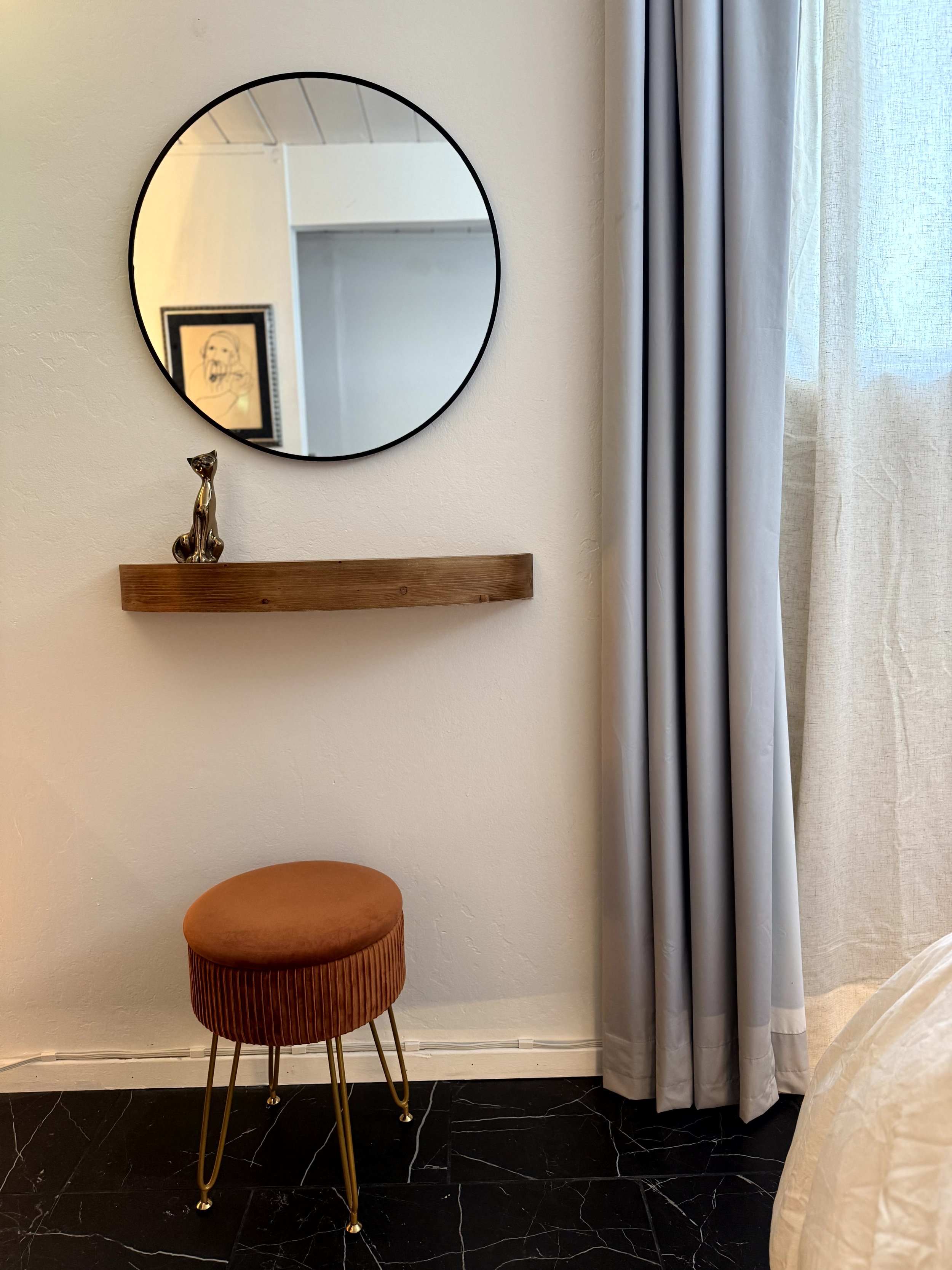 A round mirror on the wall reflects part of a room. Below the mirror, a small wooden shelf holds a decorative statuette of a sitting cat. To the right, long blue curtains cover a window. A round, orange upholstered stool with thin, gold-colored legs