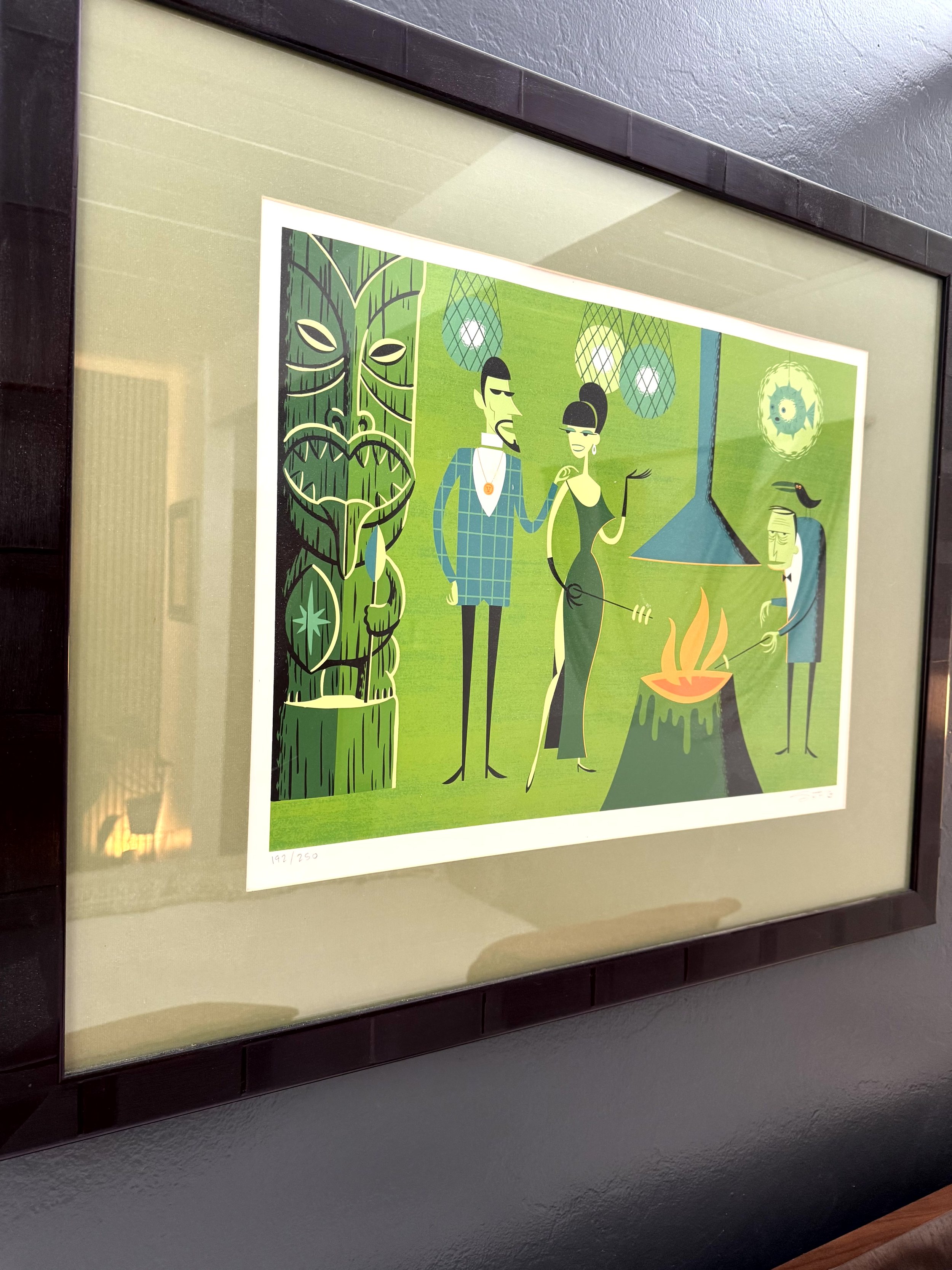Framed colorful art print depicting stylized characters having a barbecue outdoors, with a large tiki statue, a grill with flames, and hanging lanterns on a green background.