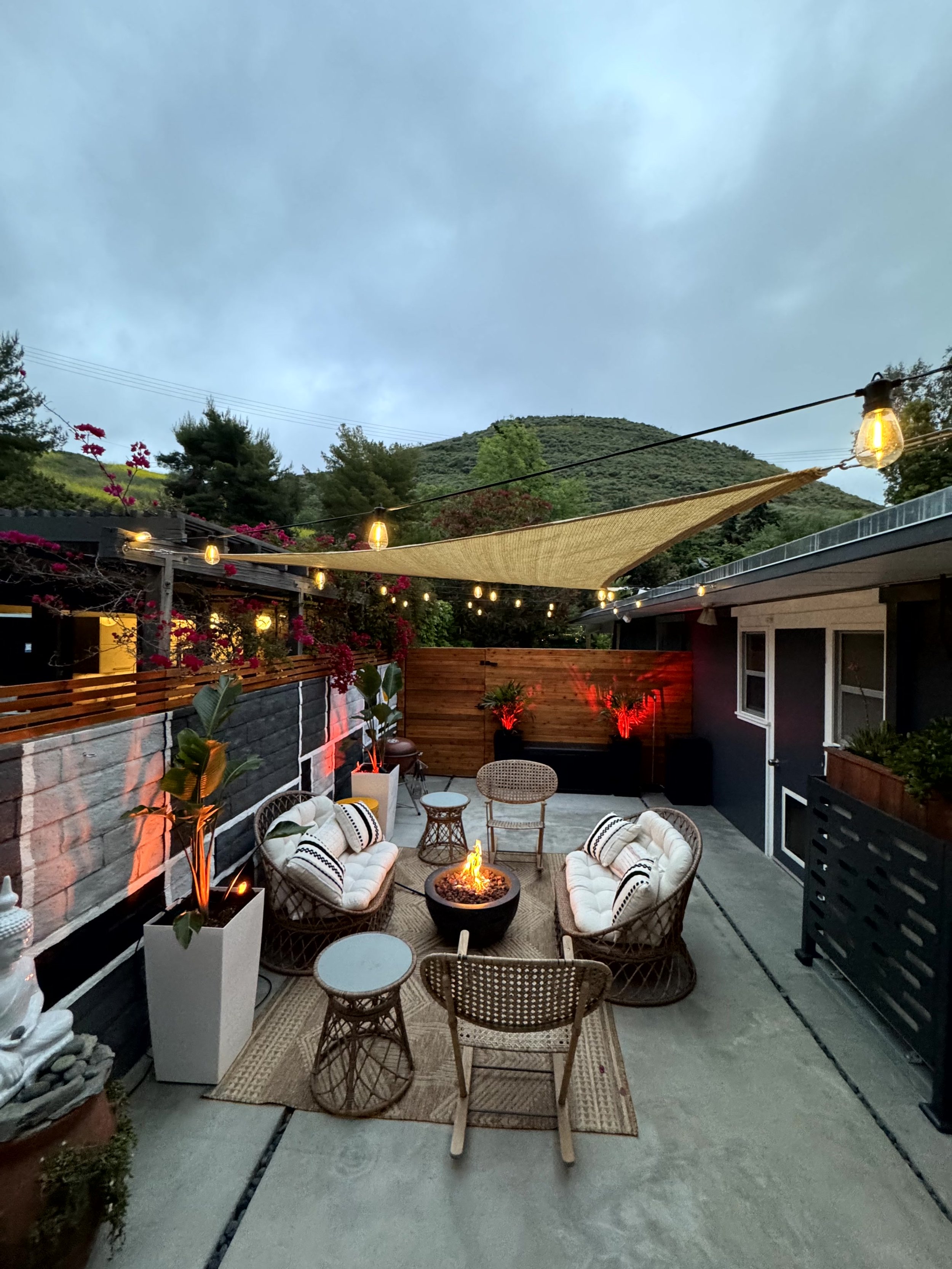 Outdoor patio with couches, chairs, and a fire pit surrounded by string lights, potted plants, and a backdrop of mountains and cloudy sky.