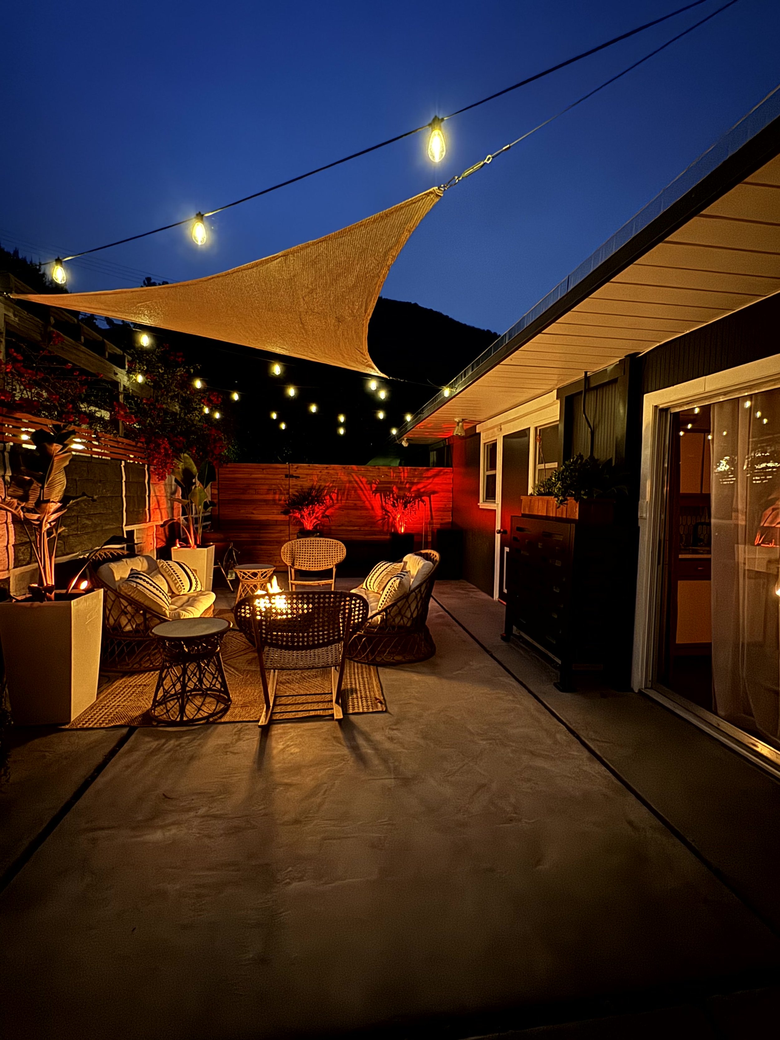 Nighttime outdoor patio with string lights, a shaded sun sail, and cozy seating area with chairs and cushions, next to a house with sliding glass doors.