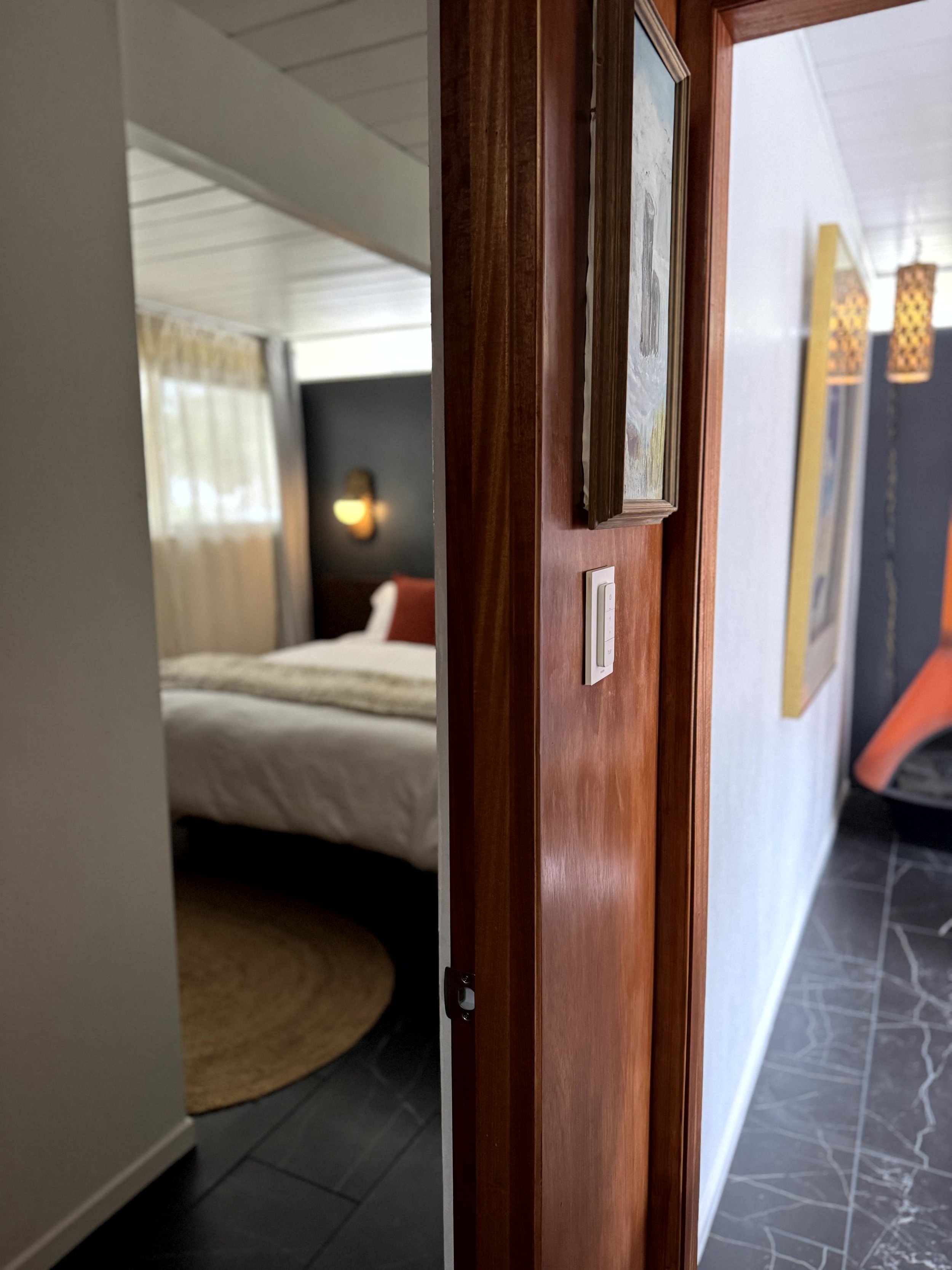 Partially open door revealing a bedroom with a bed, pillows, a wall sconce, and hanging lamps in the hallway.