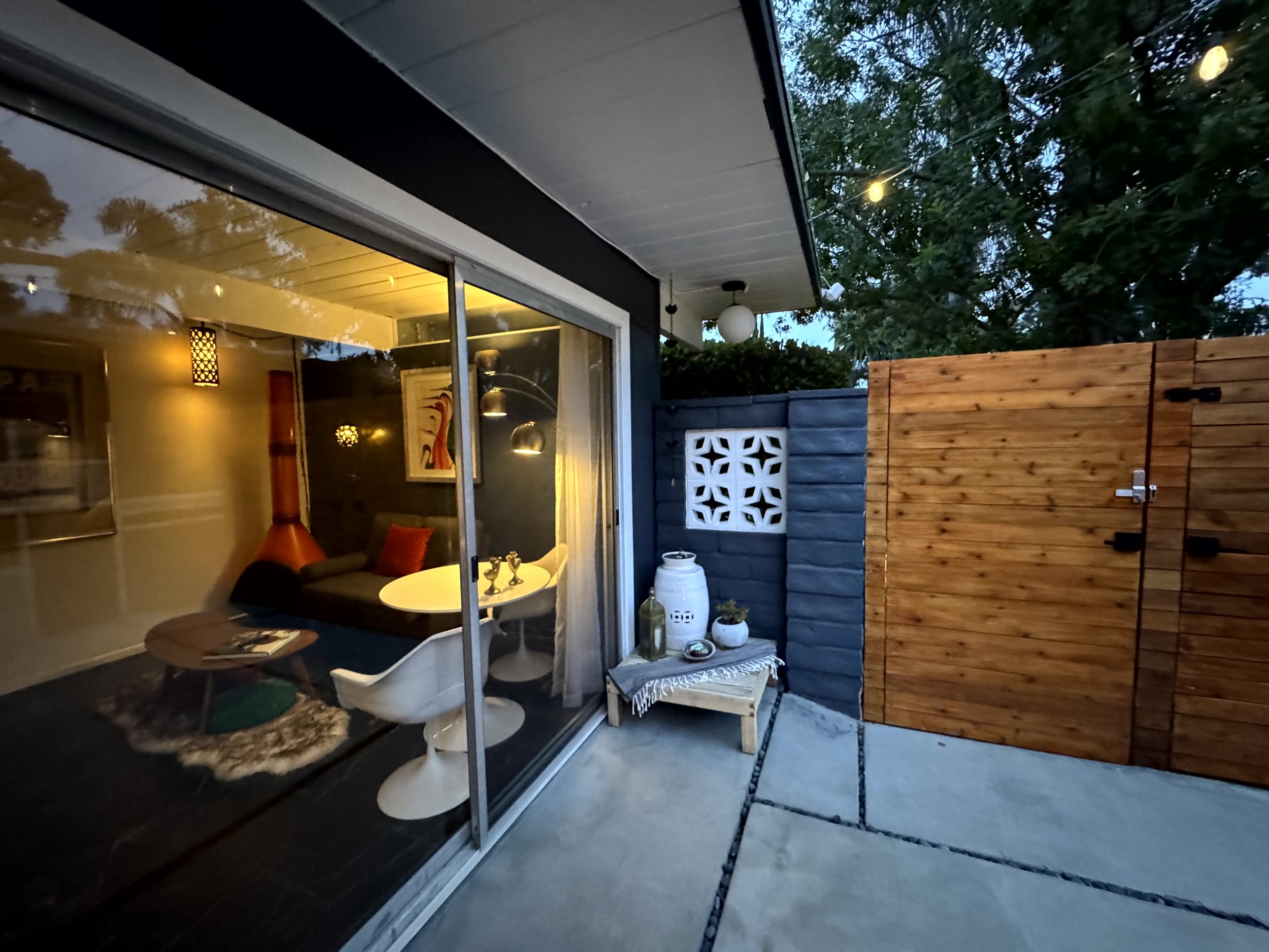 View of a modern home's outdoor patio with a wooden privacy fence, a small table with decorative items, and a large sliding glass door revealing a warmly lit living room inside.