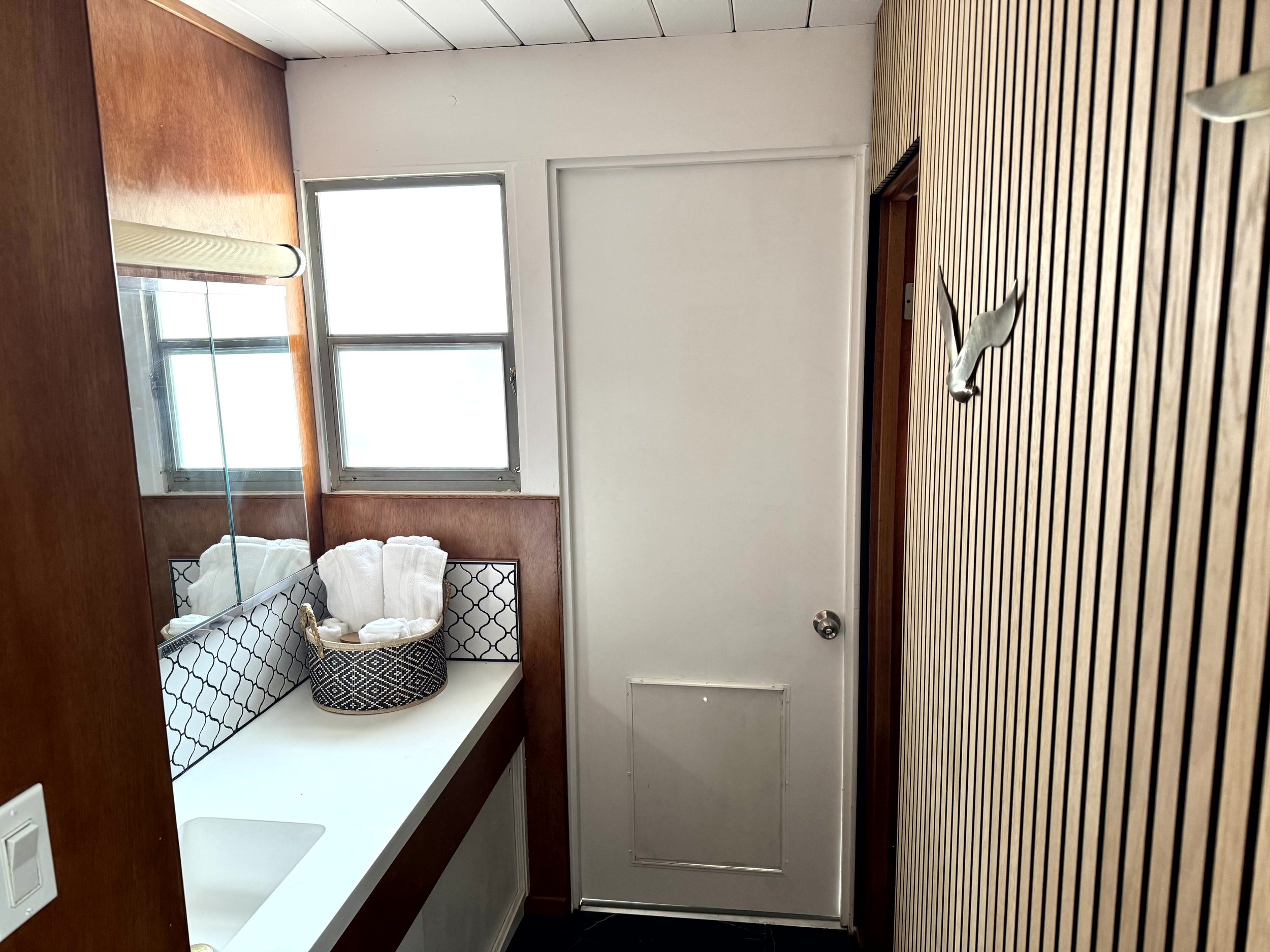 Small bathroom with wooden panel wall on the right, white door with a small pet door at the bottom, and a window above the sink with a mirror. The sink has a black and white patterned container with rolled white towels.