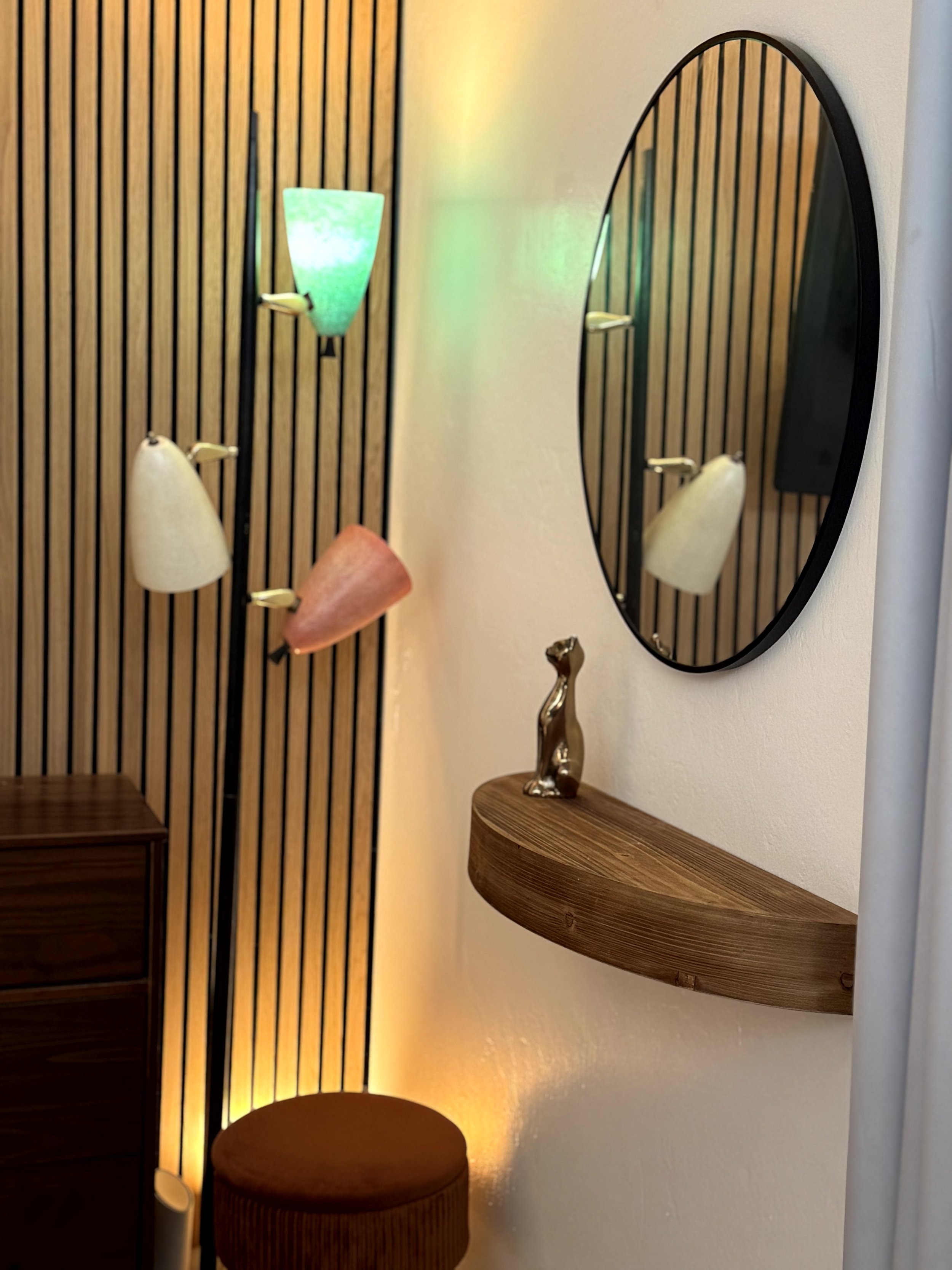 Decorative interior wall with three cone-shaped wall sconces in green, white, and red, a round mirror, a small wooden shelf with a metallic dog figurine, and a wooden cabinet, illuminated by warm and colored lighting.