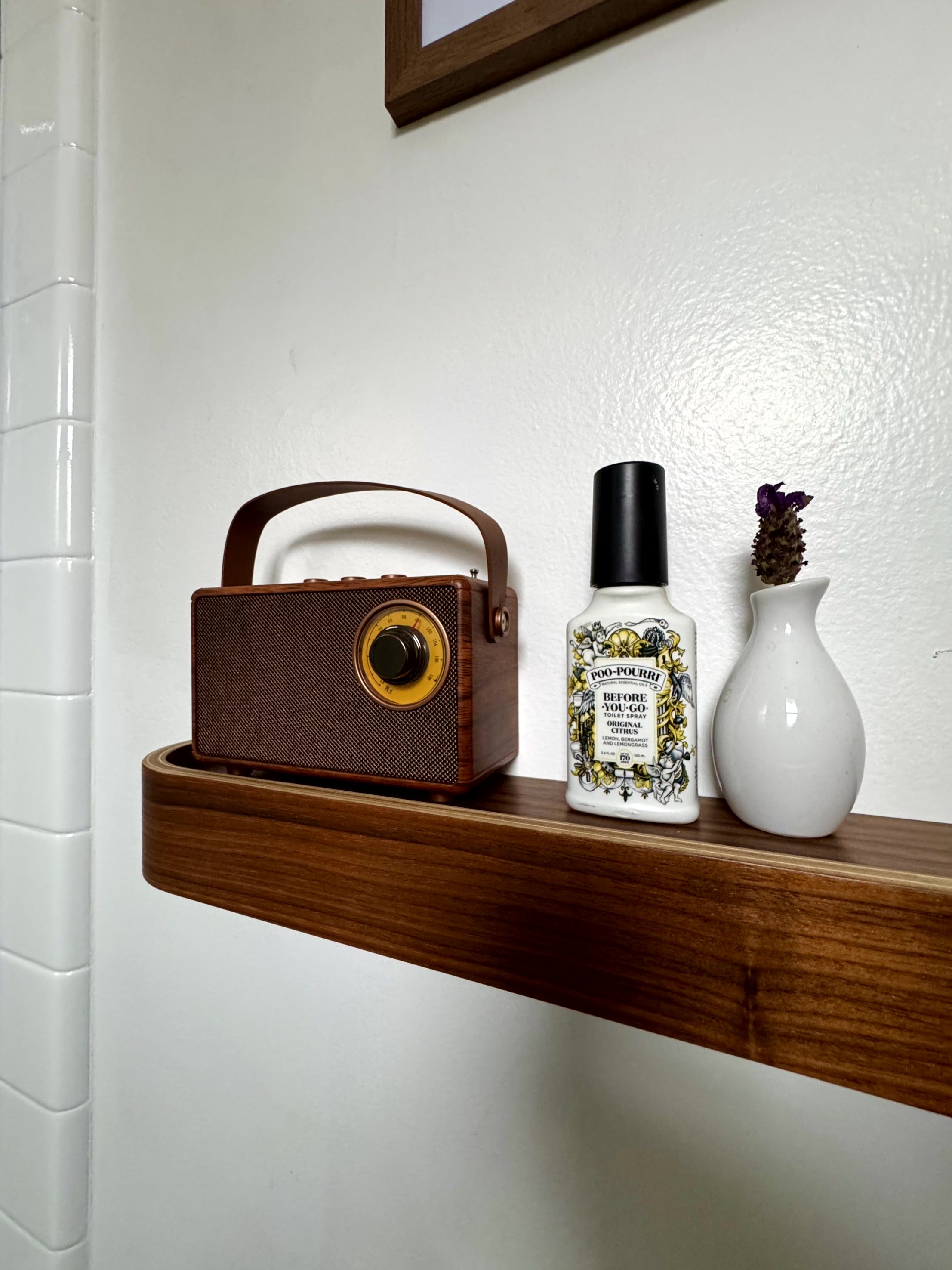 A wooden shelf holding a vintage radio, a bottle of Poo-Pourri before-you-go toilet spray, and a small white vase with a purple flower.