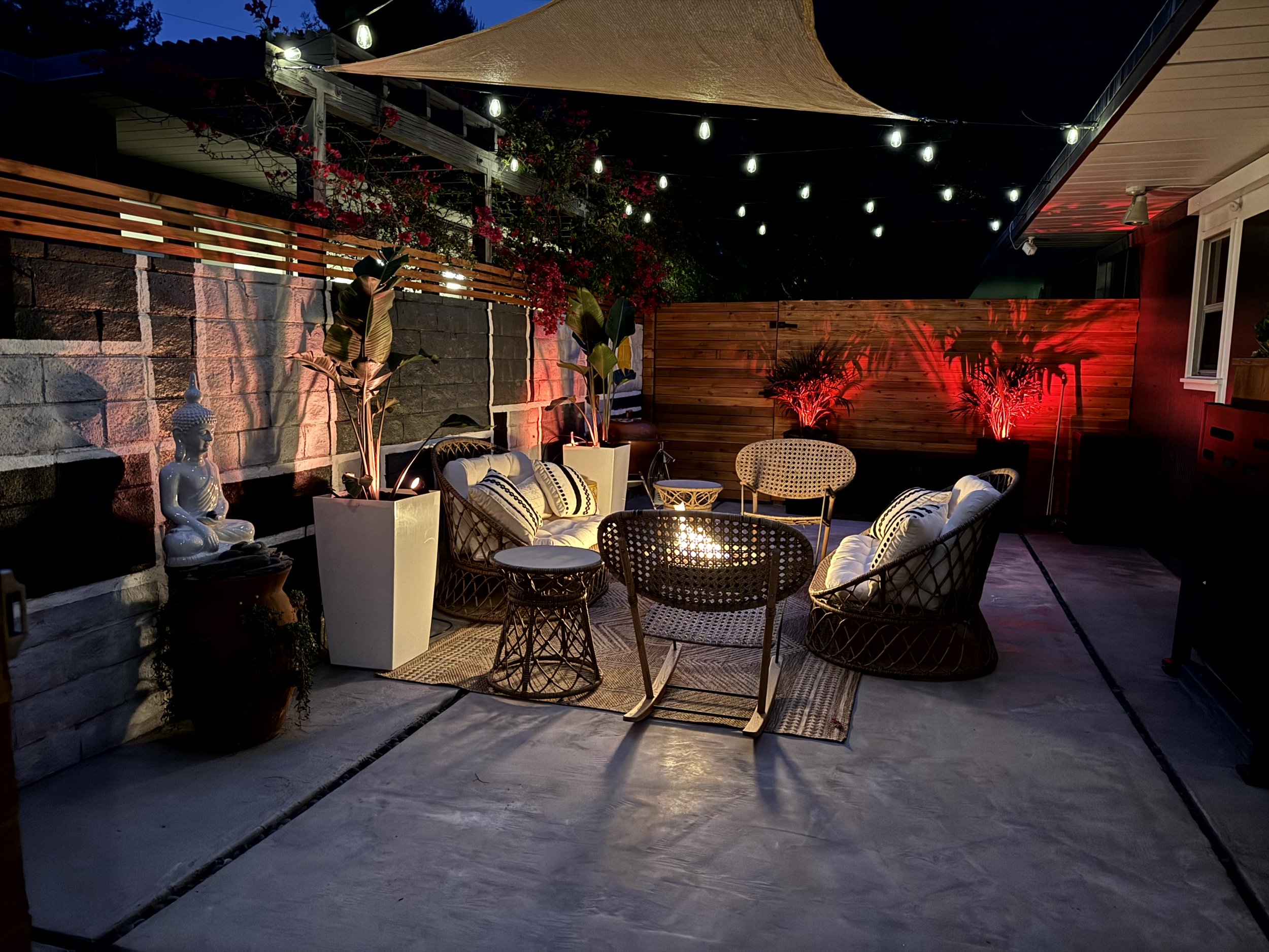 Outdoor patio at night with string lights hanging above, illuminated potted plants, sitting area with wicker chairs, cushions, and a round glass table, Buddha statue, a wooden privacy fence with red lighting, and a beige shade cloth above.