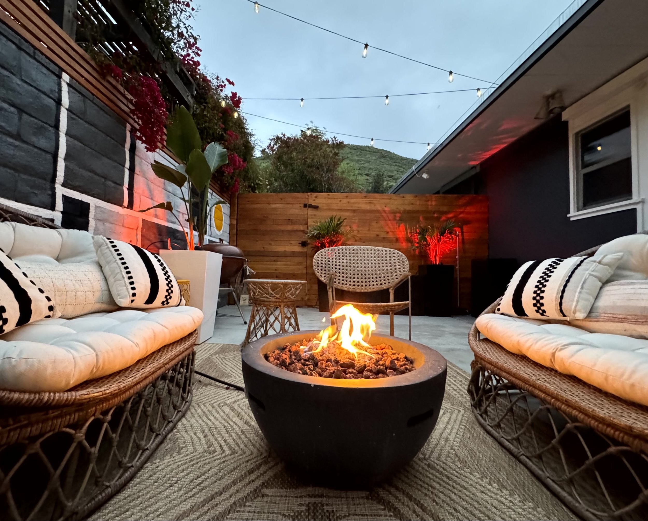 Outdoor patio with wicker couches, a fire pit, string lights, and potted plants at dusk.