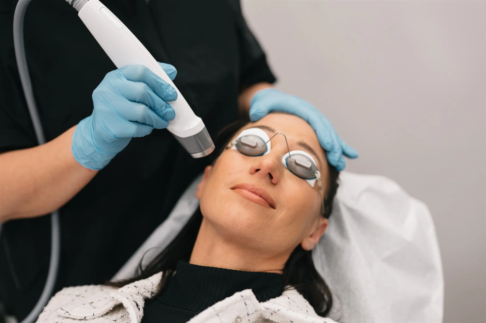 Woman receiving a laser skin treatment while lying on a medical bed with protective eye shields, a practitioner holding a laser device, and the woman smiling peacefully.