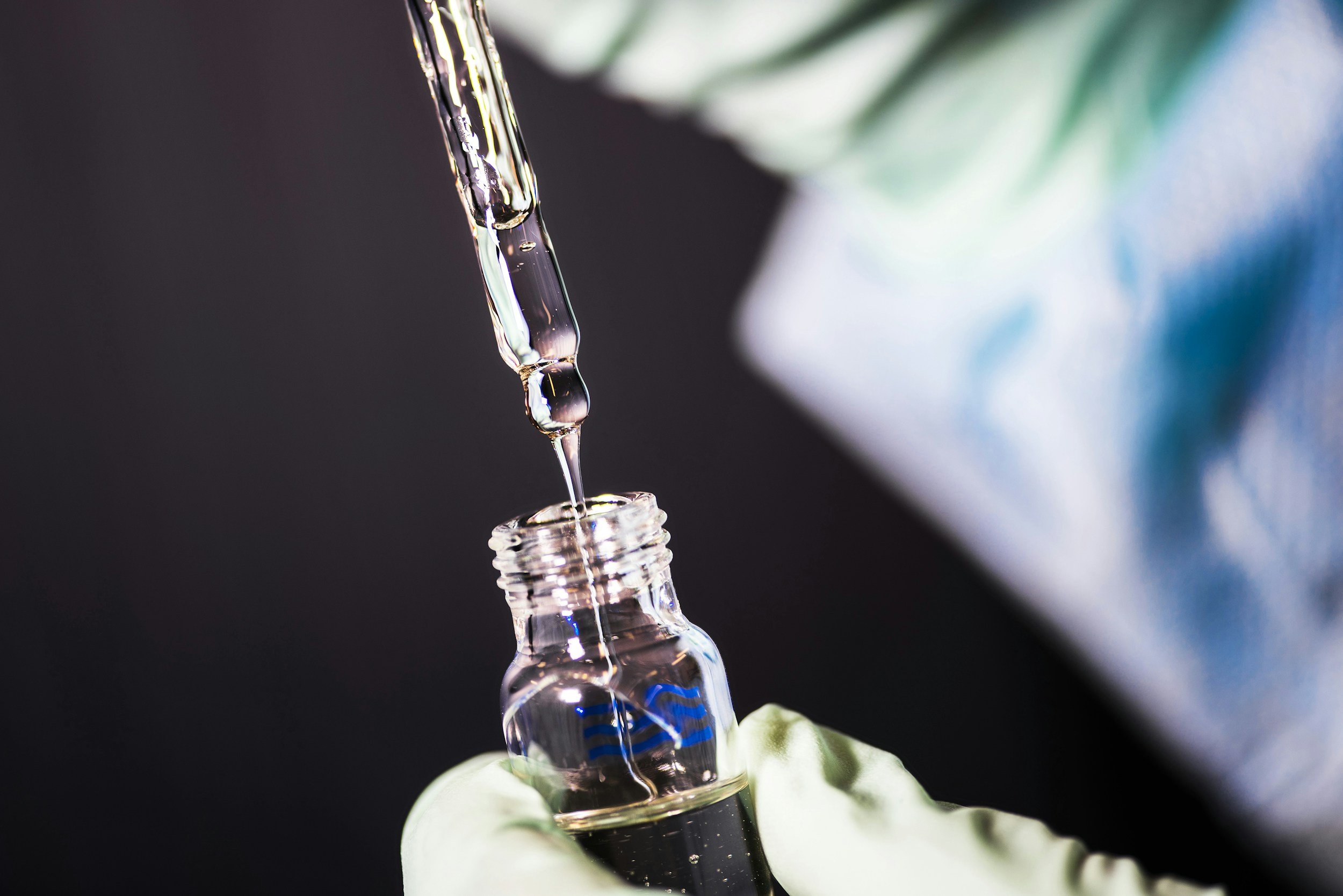 A close-up of a glass dropper releasing a drop of clear liquid into a small glass vial with a hand wearing a glove holding the vial.