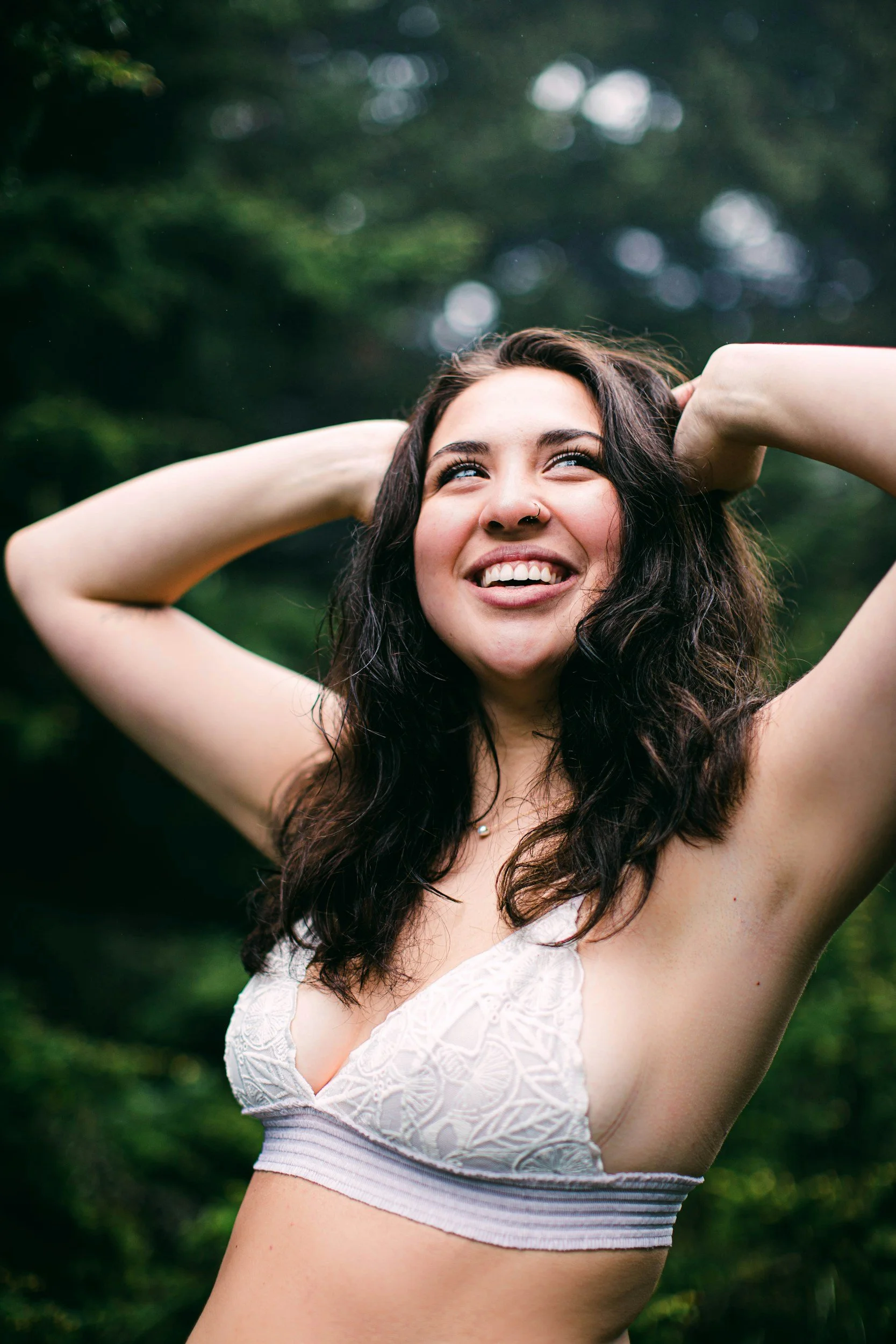 Young woman with dark curly hair smiling outdoors, wearing a white lace bralette, with hands behind her head in a natural setting.