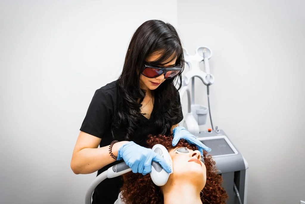 A woman with dark hair wearing protective glasses and gloves performing a procedure on a patient with curly red hair lying back with eye protection on, in a medical clinic setting.