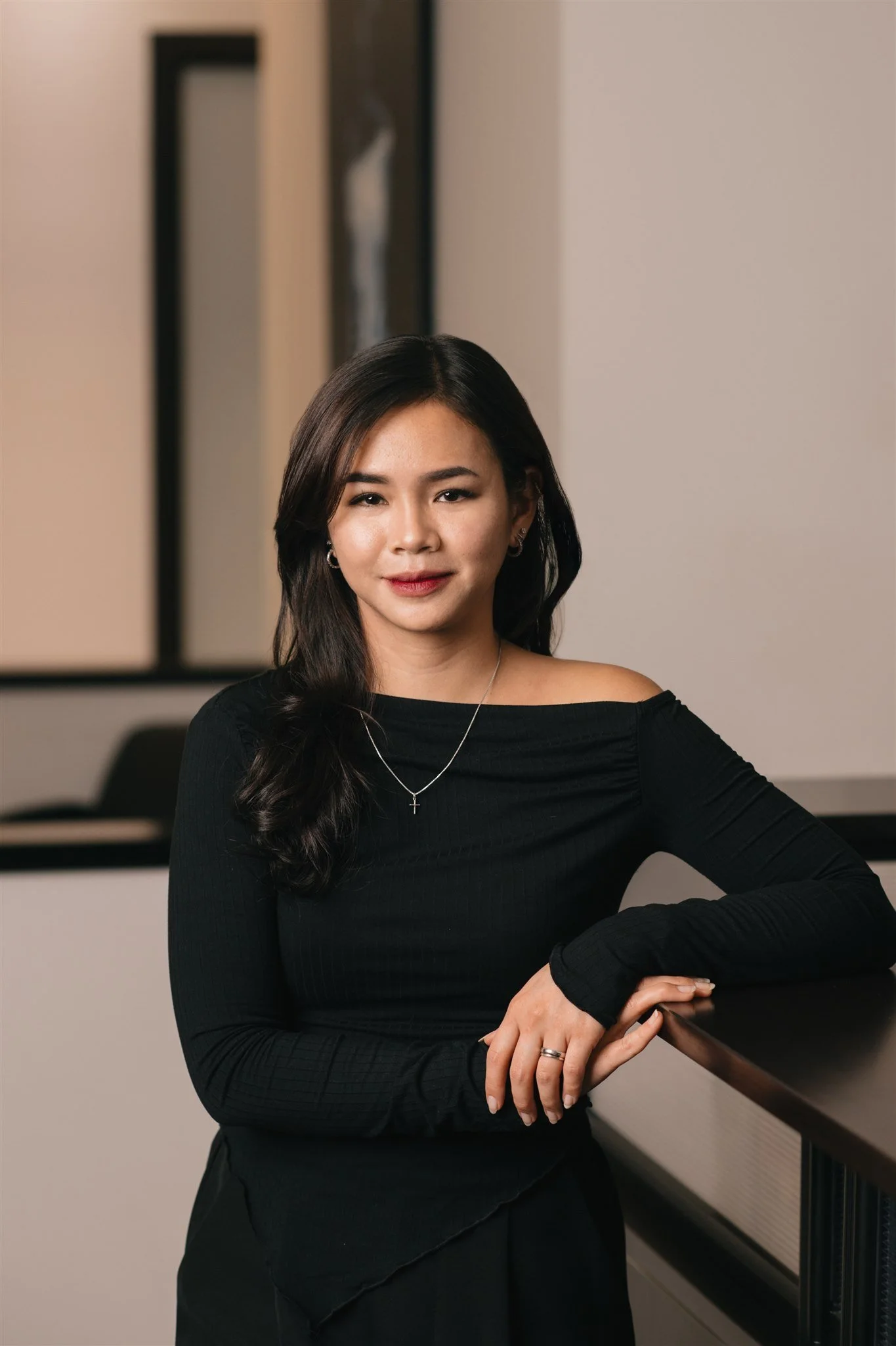 A woman with long dark hair and wearing a black off-shoulder top, standing at a counter with her arms resting on it, in an indoor setting with modern decor.
