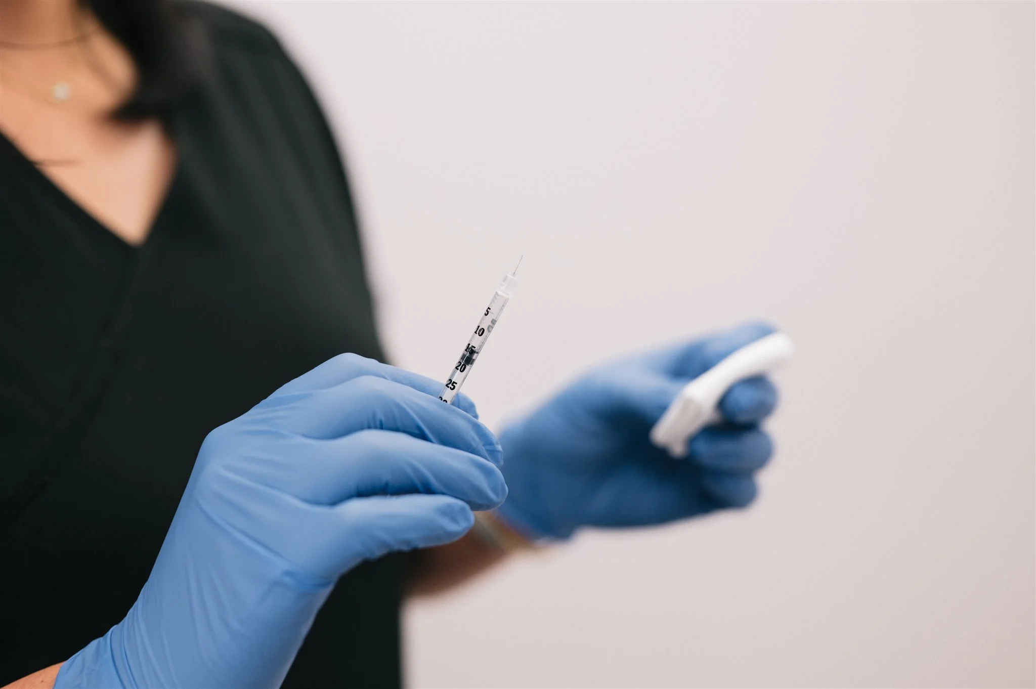 Person wearing blue gloves holding a syringe and a cotton pad, preparing to administer an injection against a plain background.