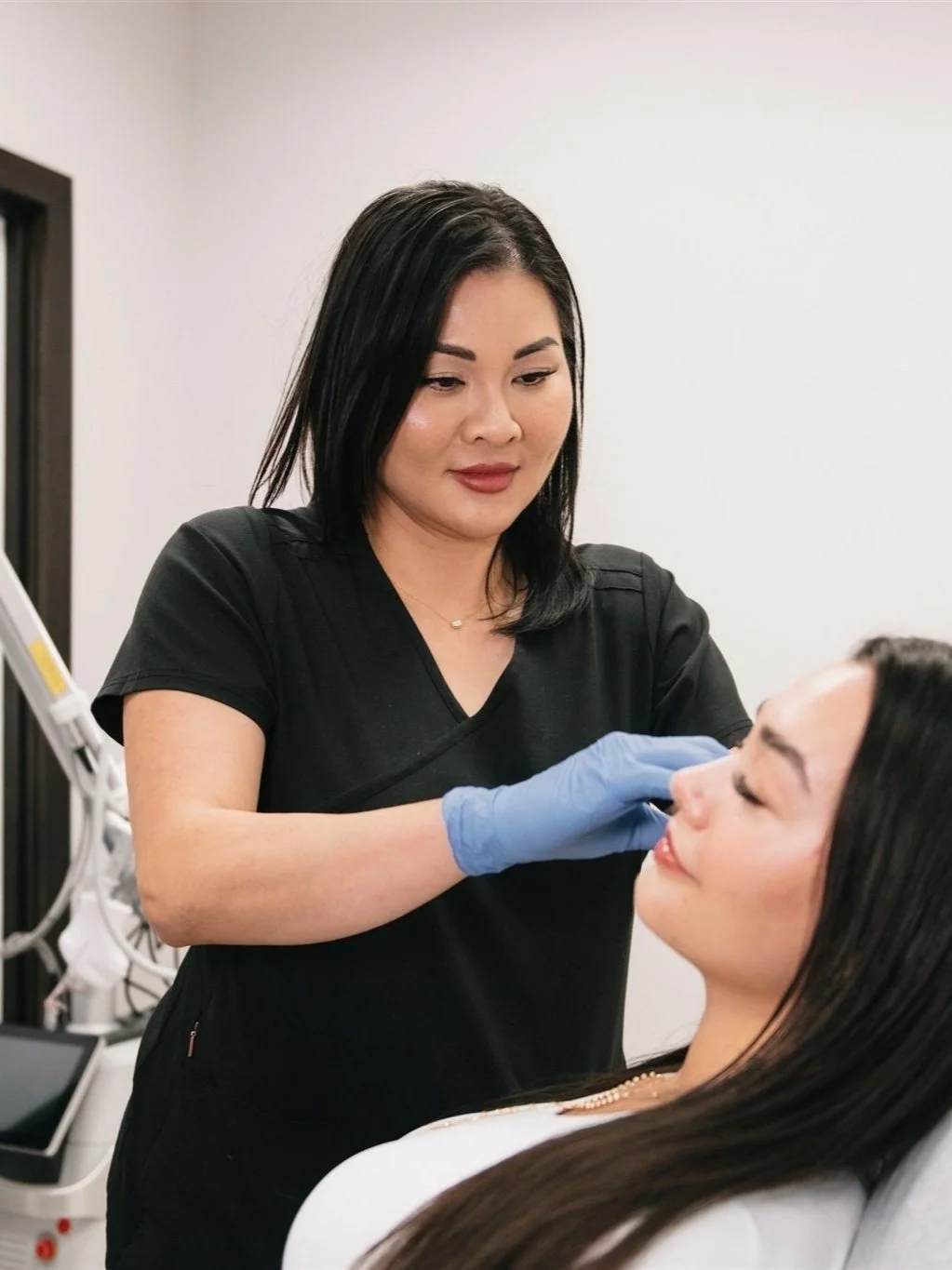 A woman with black hair and a black uniform applying makeup or skincare on a seated woman with long dark hair in a clinical or spa setting.