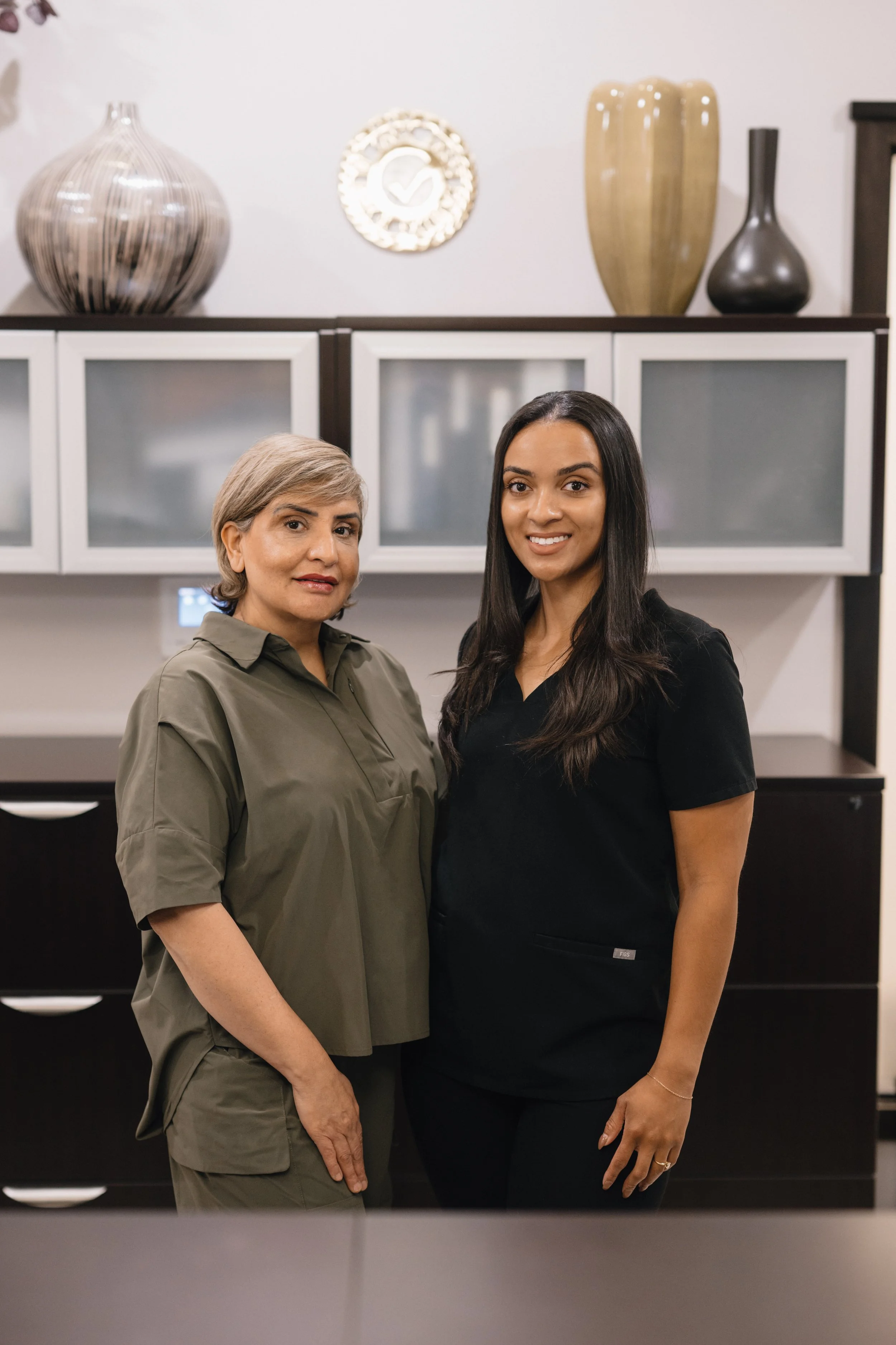 Two women, one older with gray hair and one younger with long dark hair, standing side by side in a modern room with decorative vases and wall art behind them.