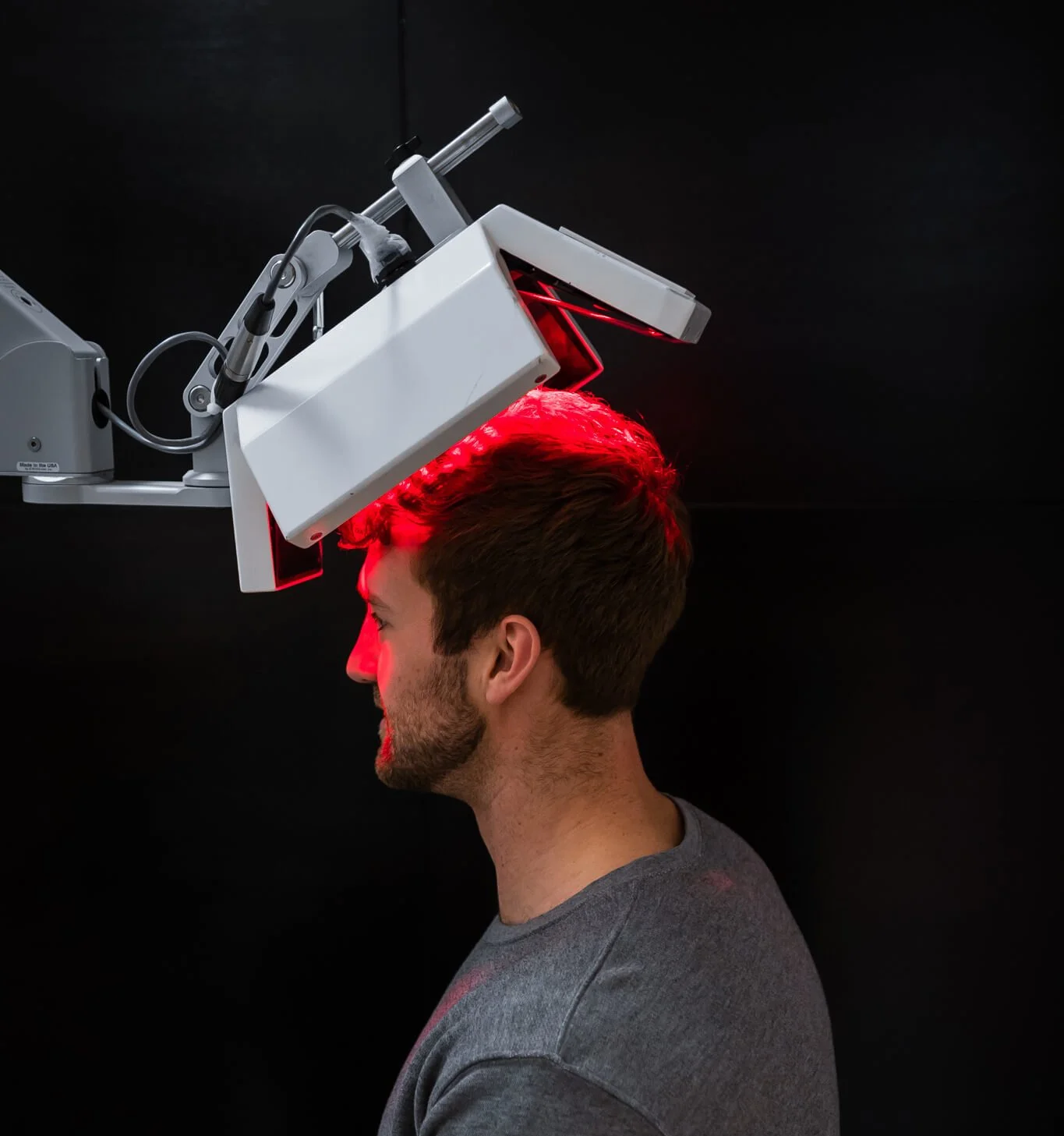 A man is seated with a red laser device above his head, directed at his scalp, possibly for a medical or cosmetic treatment, against a black background.