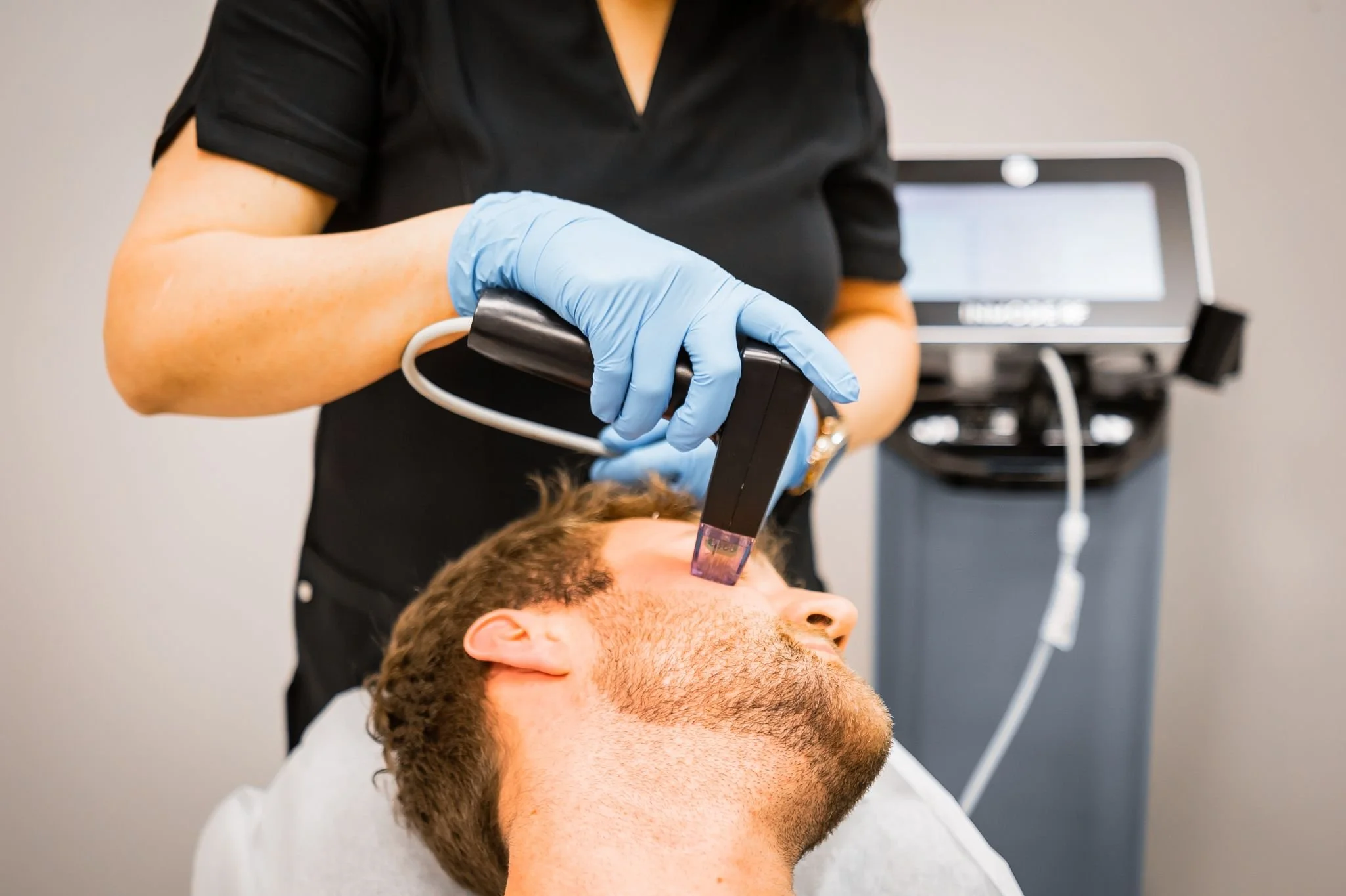 A healthcare professional in black uniform and blue gloves applying a laser treatment to a man's forehead in a clinical setting.