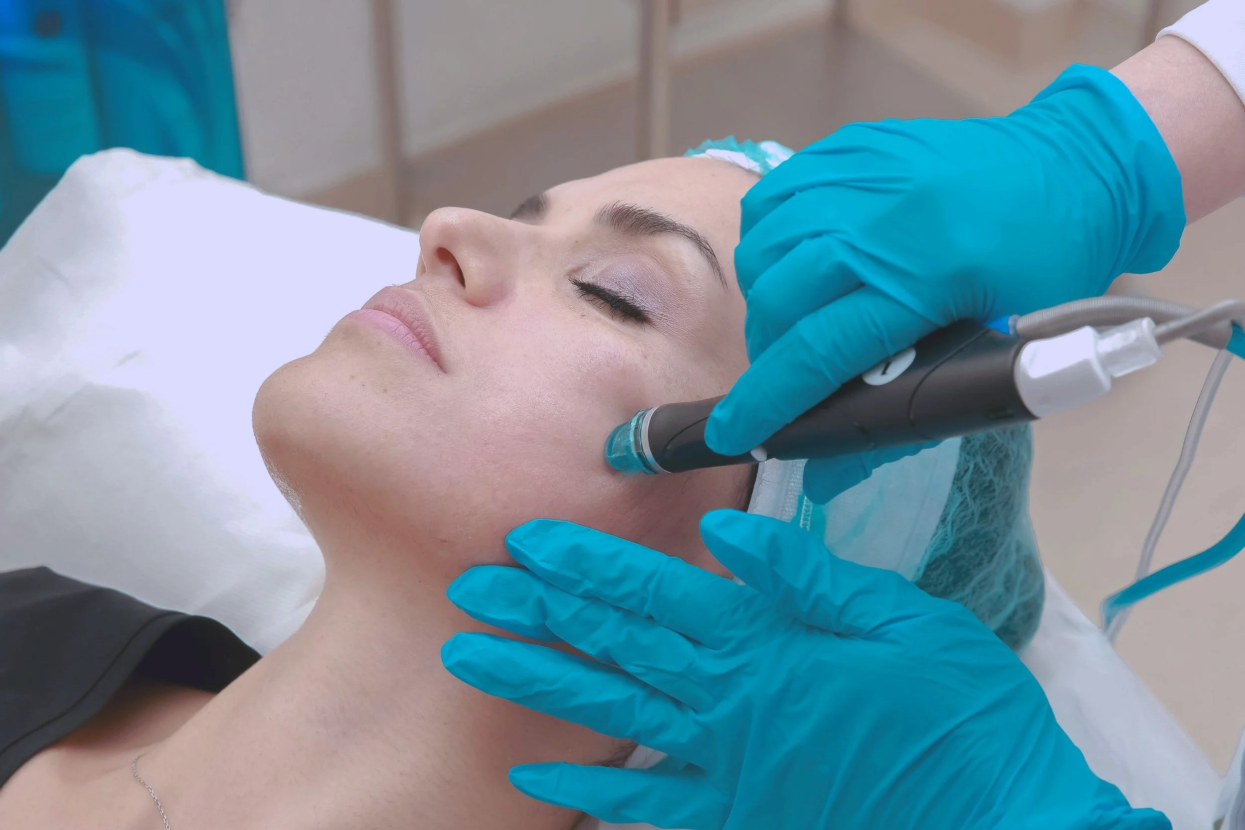 A woman lying on a medical bed receiving a facial treatment with a specialized device from a healthcare professional wearing blue gloves.